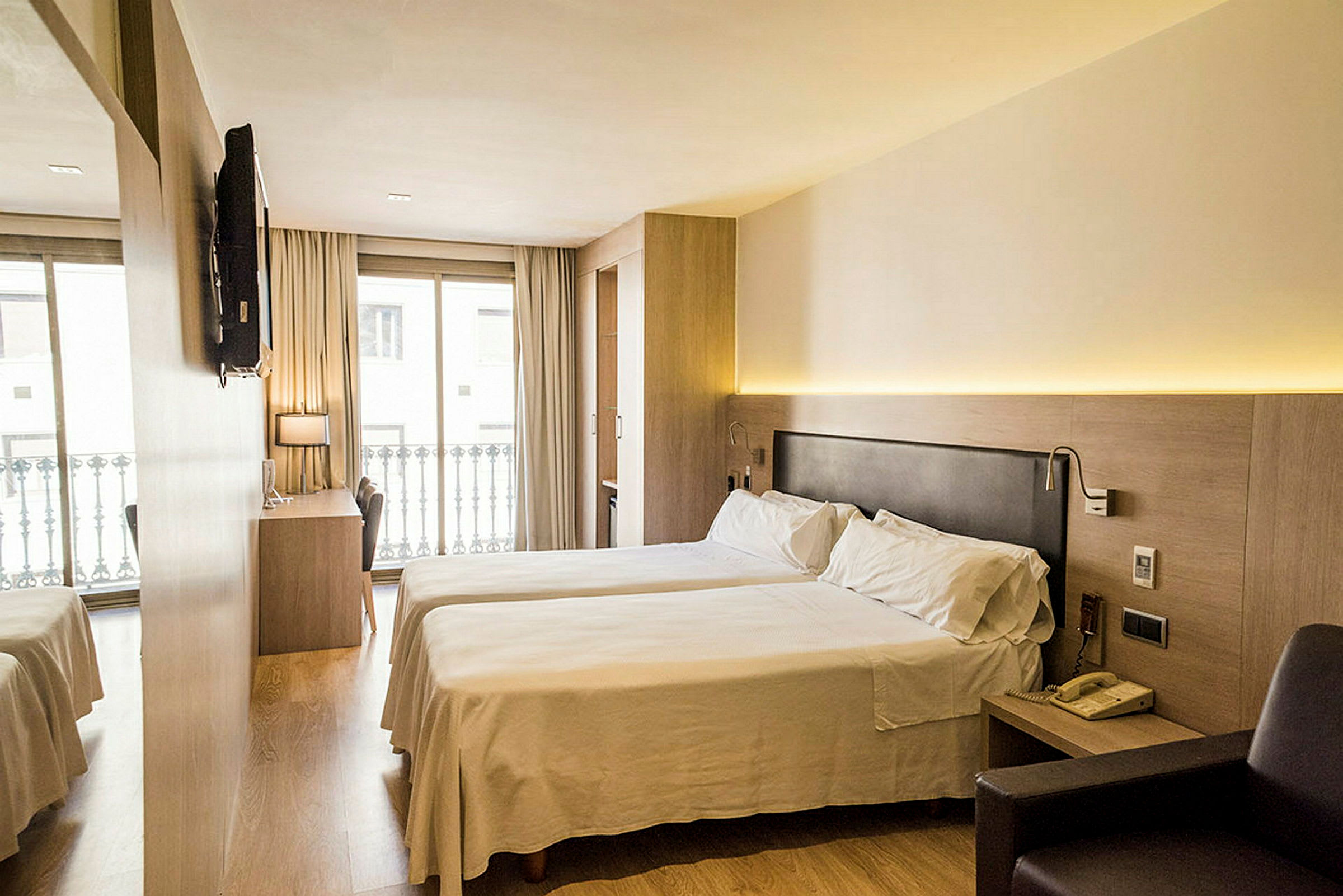 Ett hotellrum med två enkelsängar, ett skrivbord och en stol framför en stor fönster med balkongräcke. Ett TV är monterad på väggen. (AI-genererad text)