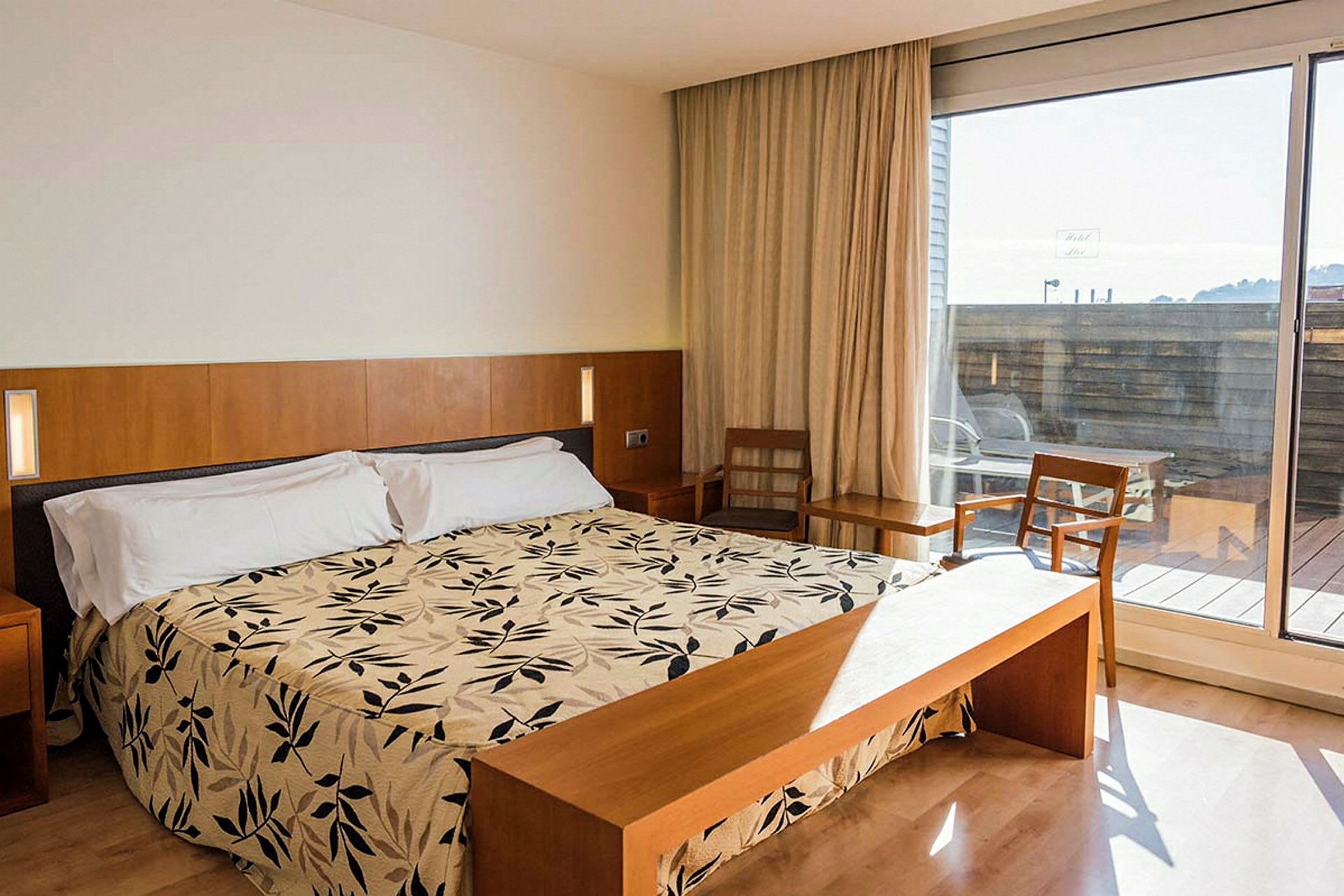 Ett hotellrum med en dubbelsäng med mönstrat täcke, trämöbler och stora glasdörrar ut mot en terrass. (AI-genererad text)