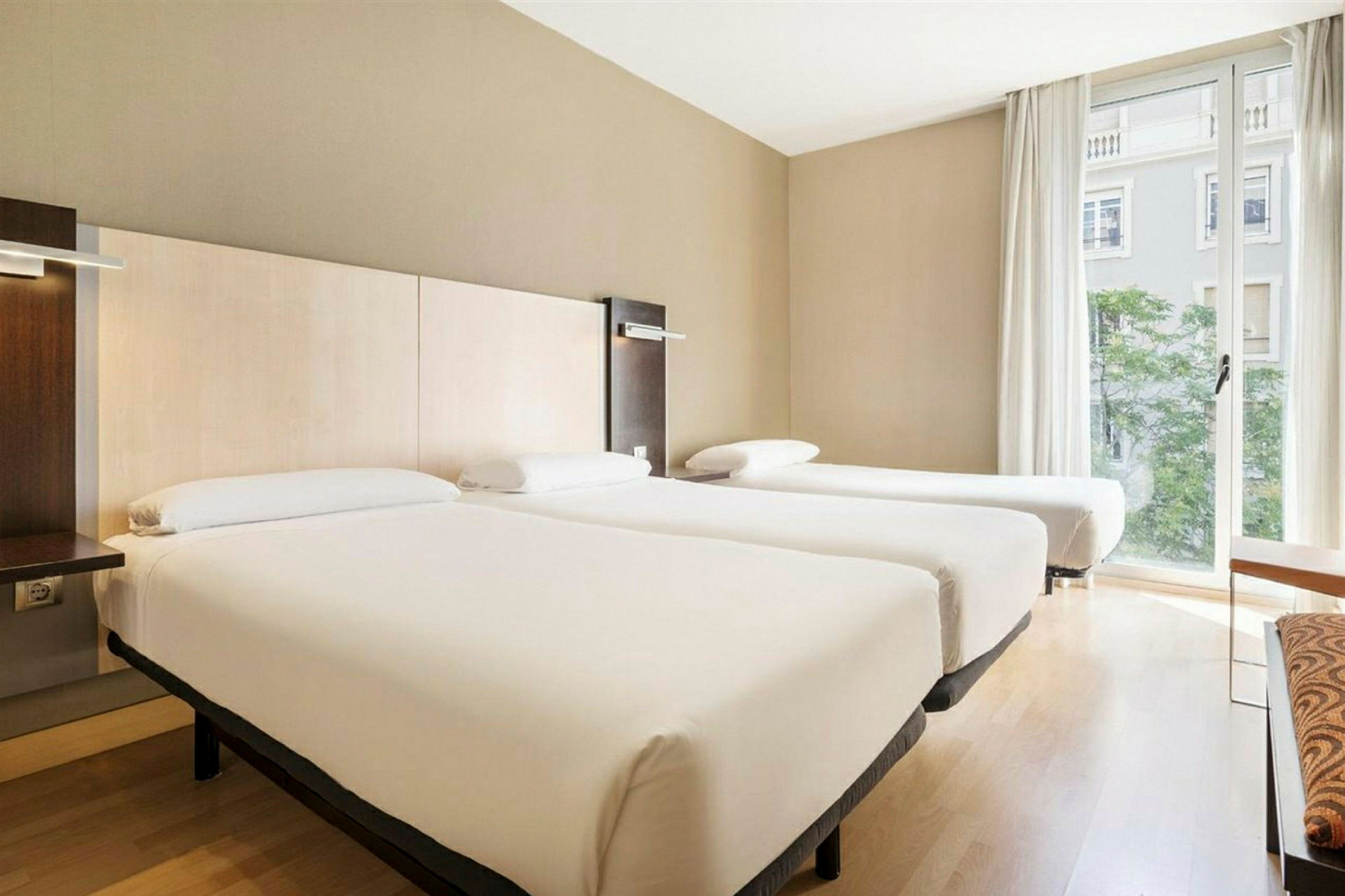 Ett hotellrum med tre enkelsängar bredvid varandra, ljust inrett och med ett fönster som vetter mot en byggnad. (AI-genererad text)