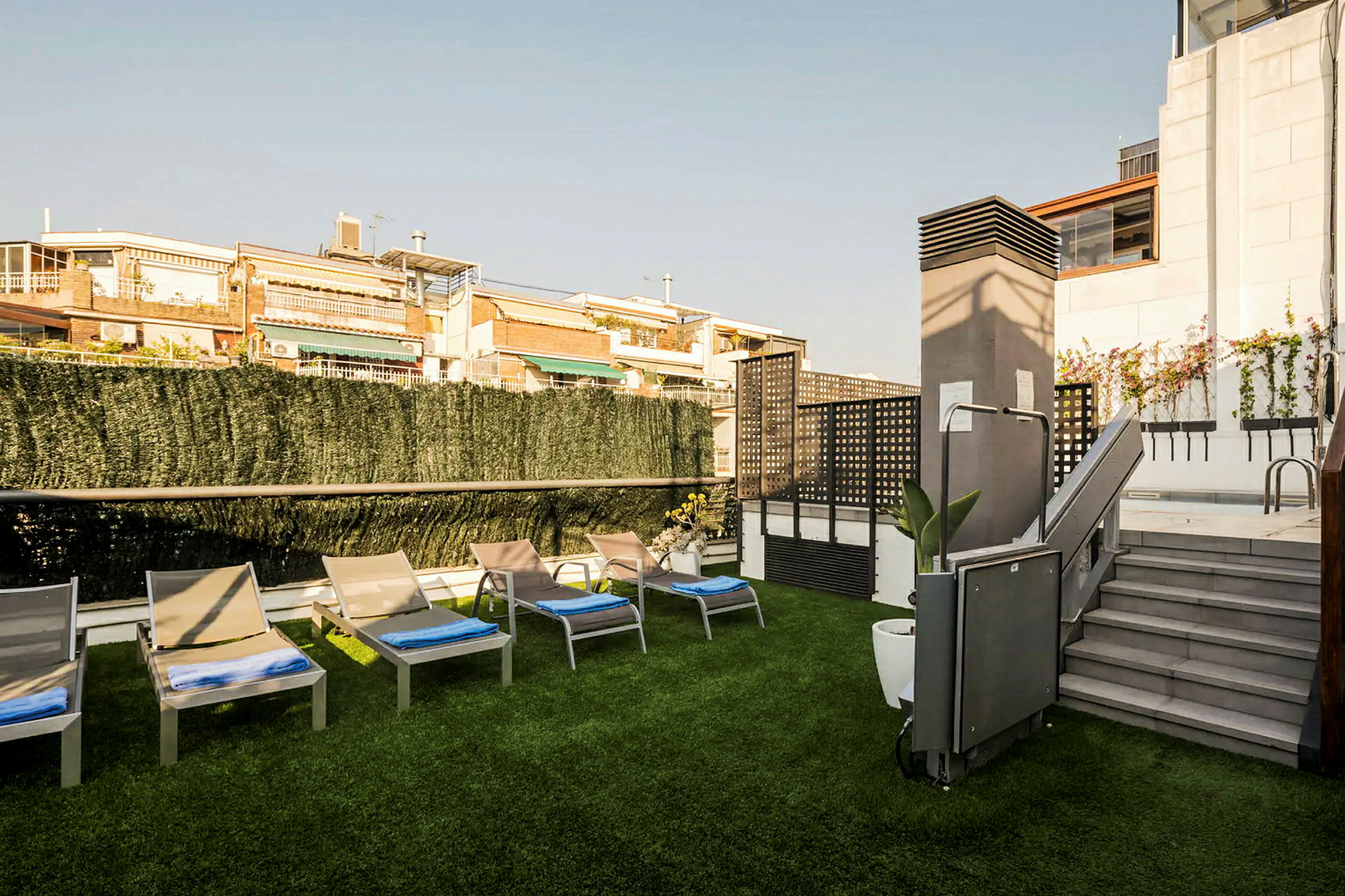 Bild av en solterrass med flera solstolar på ett område med konstgräs, omgiven av gröna häckar och byggnader i bakgrunden. (AI-genererad text)