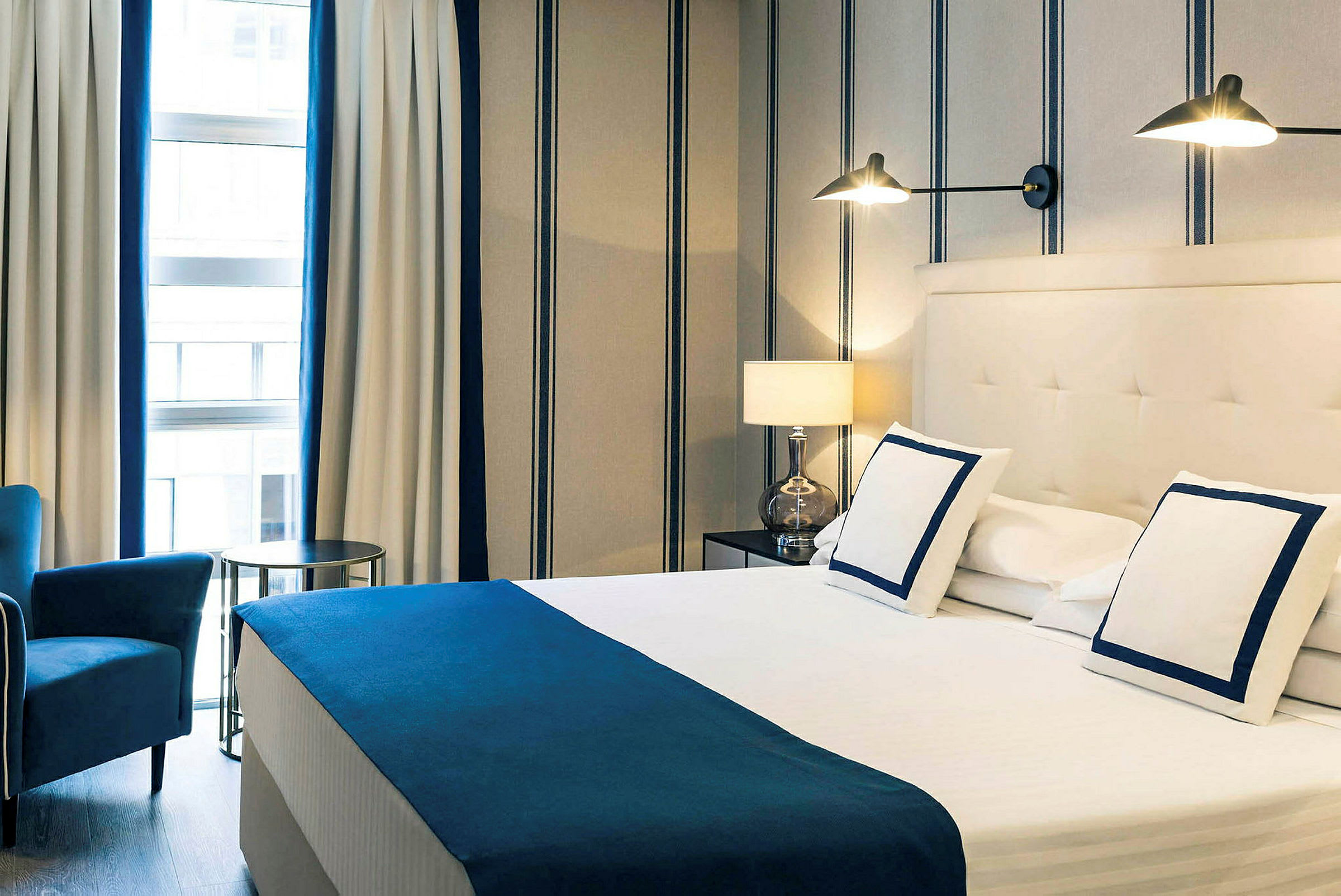 Ett hotellrum med en dubbelsäng, vita sängkläder med blå detaljer, en armstol, och randiga väggar samt gardiner. (AI-genererad text)