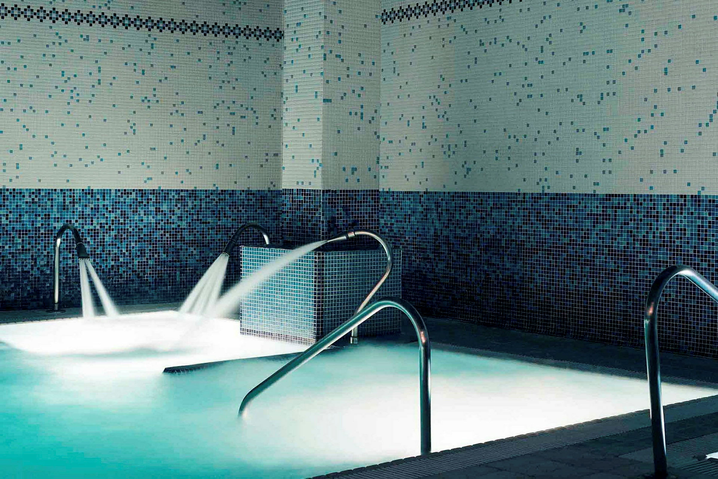 Inomhuspool med blå mosaikväggar och metallräcken, vatten sprutar ur flera uttag. (AI-genererad text)