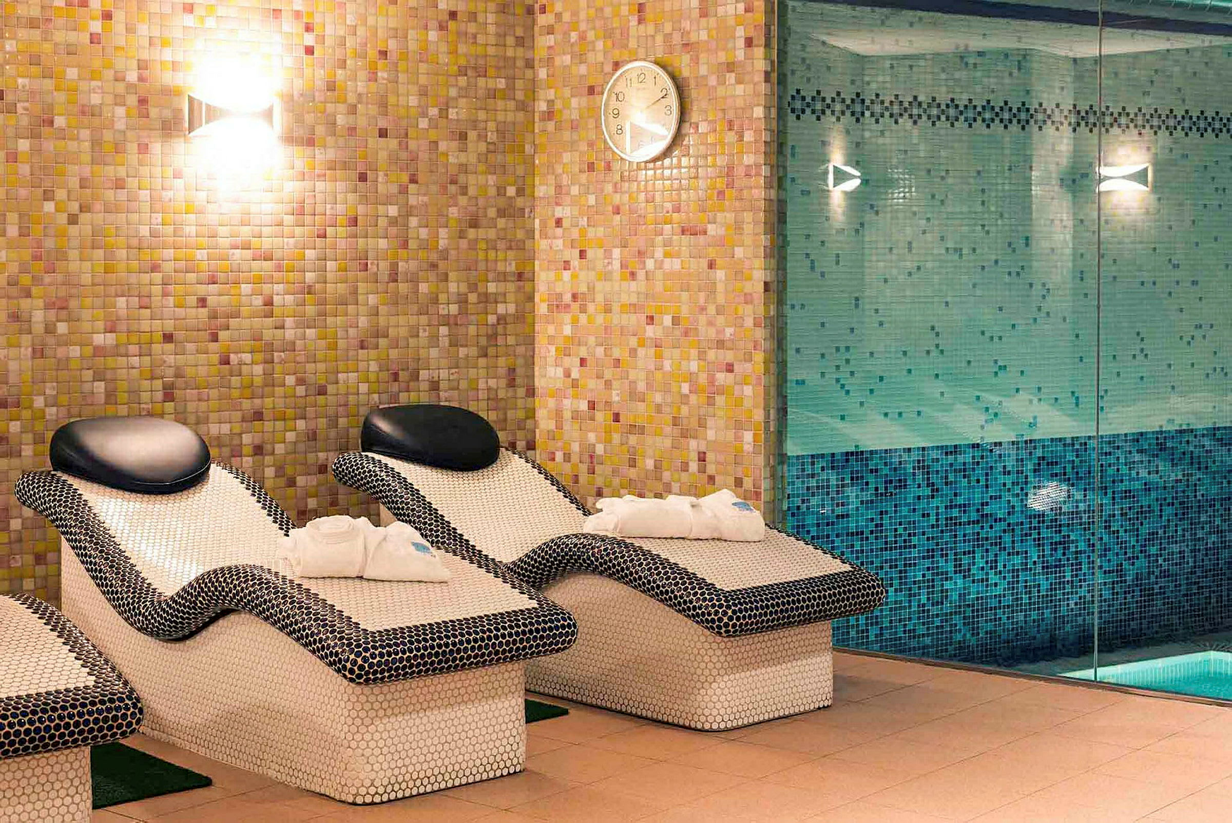 Bild av ett avkopplingsområde med mosaikklädda vilstolar och en pool bakom glasvägg. (AI-genererad text)