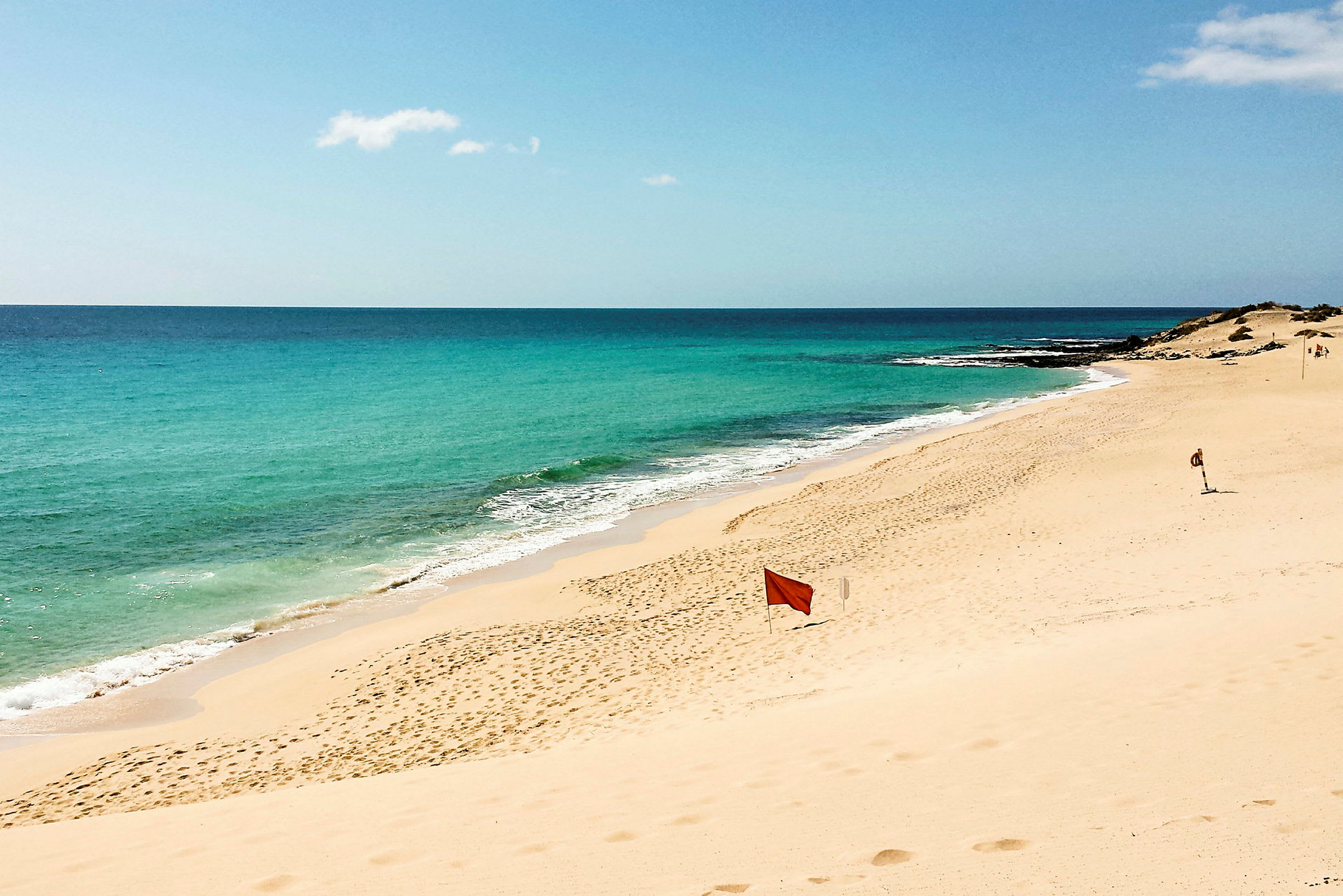 En sandstrand med turkost vatten och blå himmel. En röd varningsflagga står på stranden. (AI-genererad text)