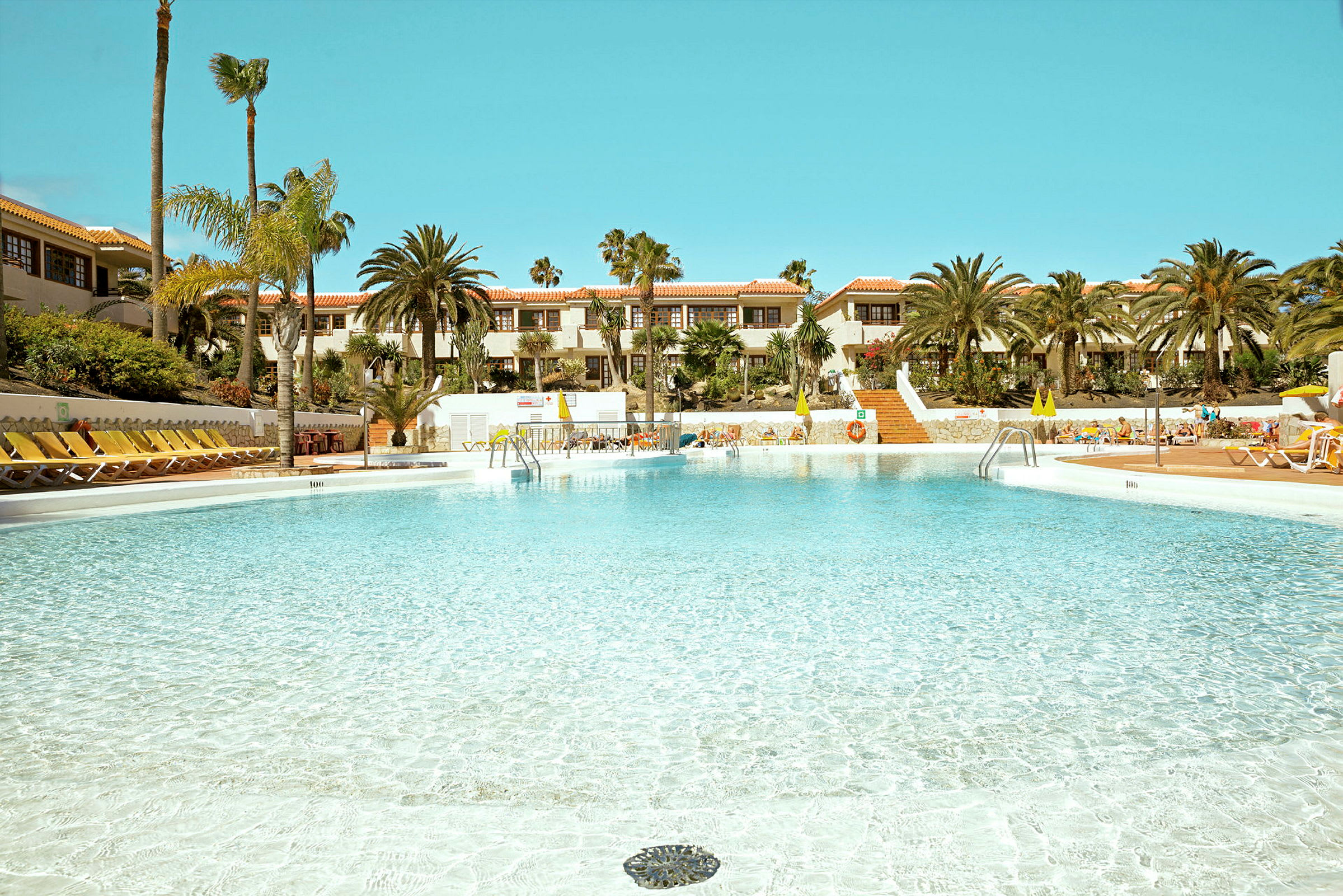 En stor pool vid ett hotellkomplex med solstolar och palmer. (AI-genererad text)