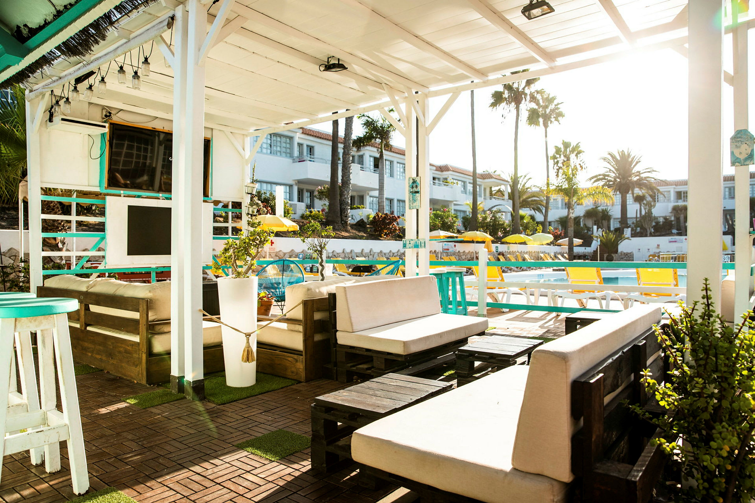 En utomhuslounge under en vit pergola med möbler och växter, med utsikt över en pool och omgivande byggnader. (AI-genererad text)