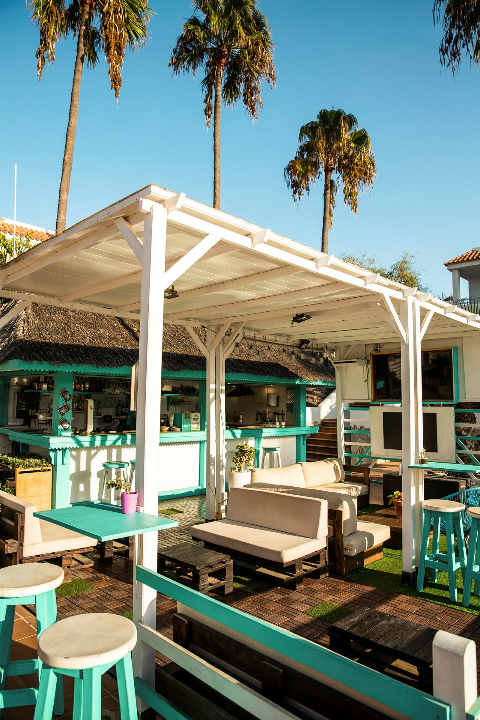 En utomhuslounge med soffor och bord under en vit träpergola, omgiven av palmer och en bar. (AI-genererad text)