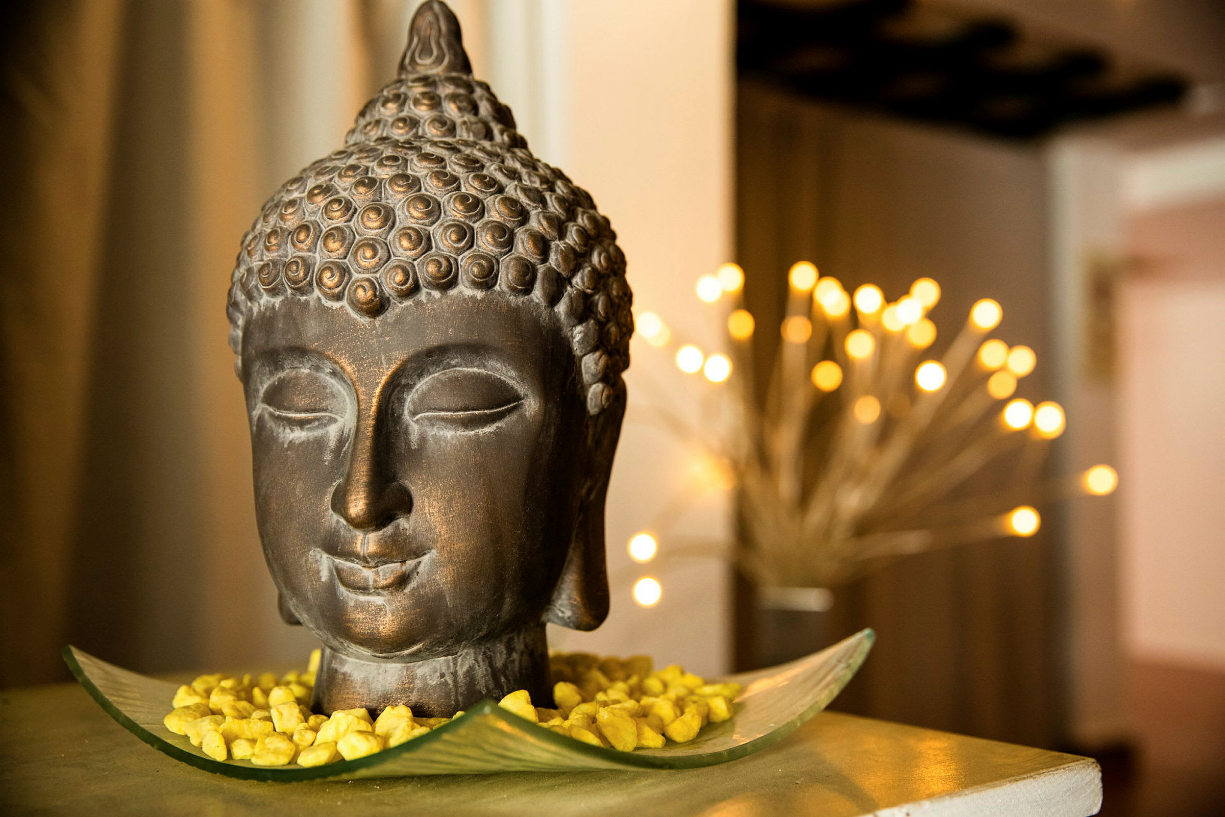 En dekorativ Buddha-figur placerad på ett fat med guldfärgade stenar. Mjukt ljusskimmer i bakgrunden. (AI-genererad text)