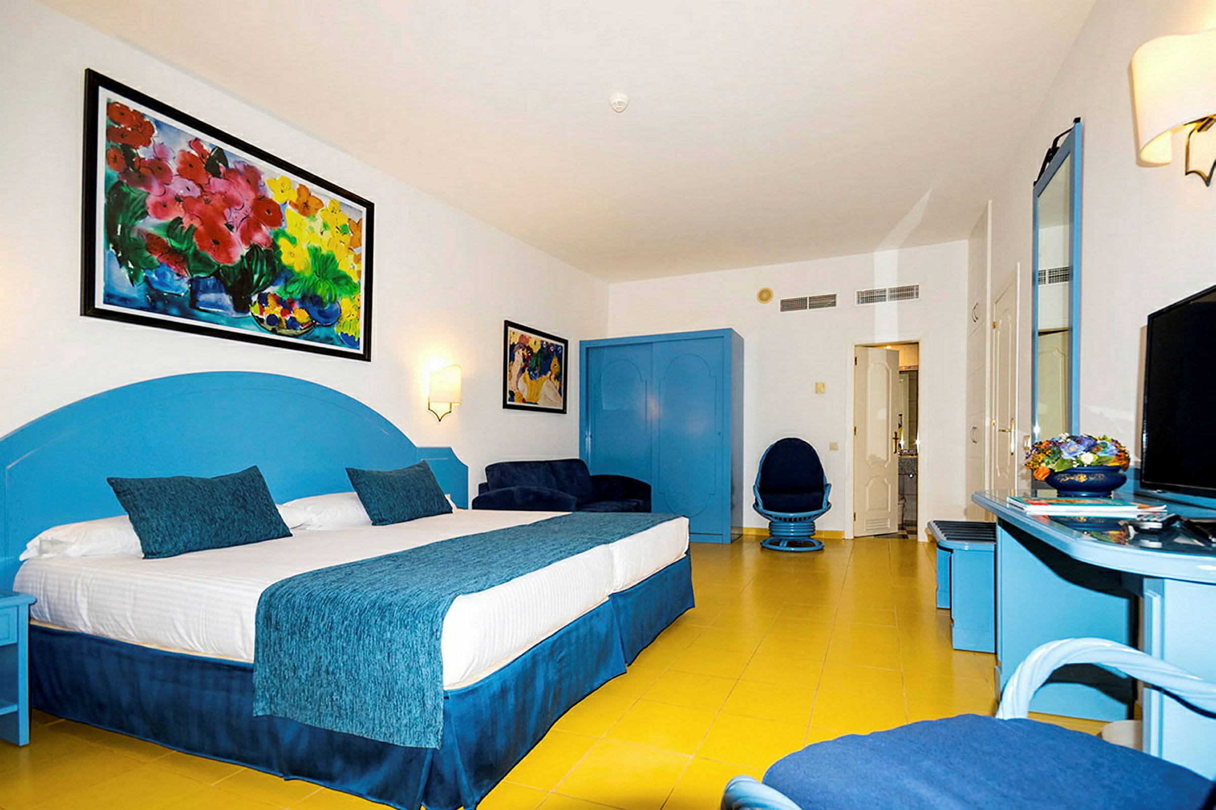 Ett hotellrum med en stor dubbelsäng med blå detaljer, färgglada väggmålningar, en blå byrå och fåtöljer. (AI-genererad text)
