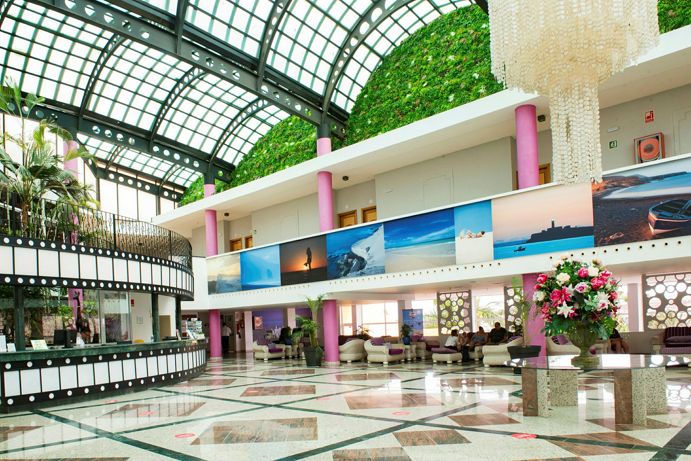 En rymlig hotellobby med en glastäckt takstruktur och gröna väggar. En stor ljuskrona och färgstarka växter står i fokus. (AI-genererad text)