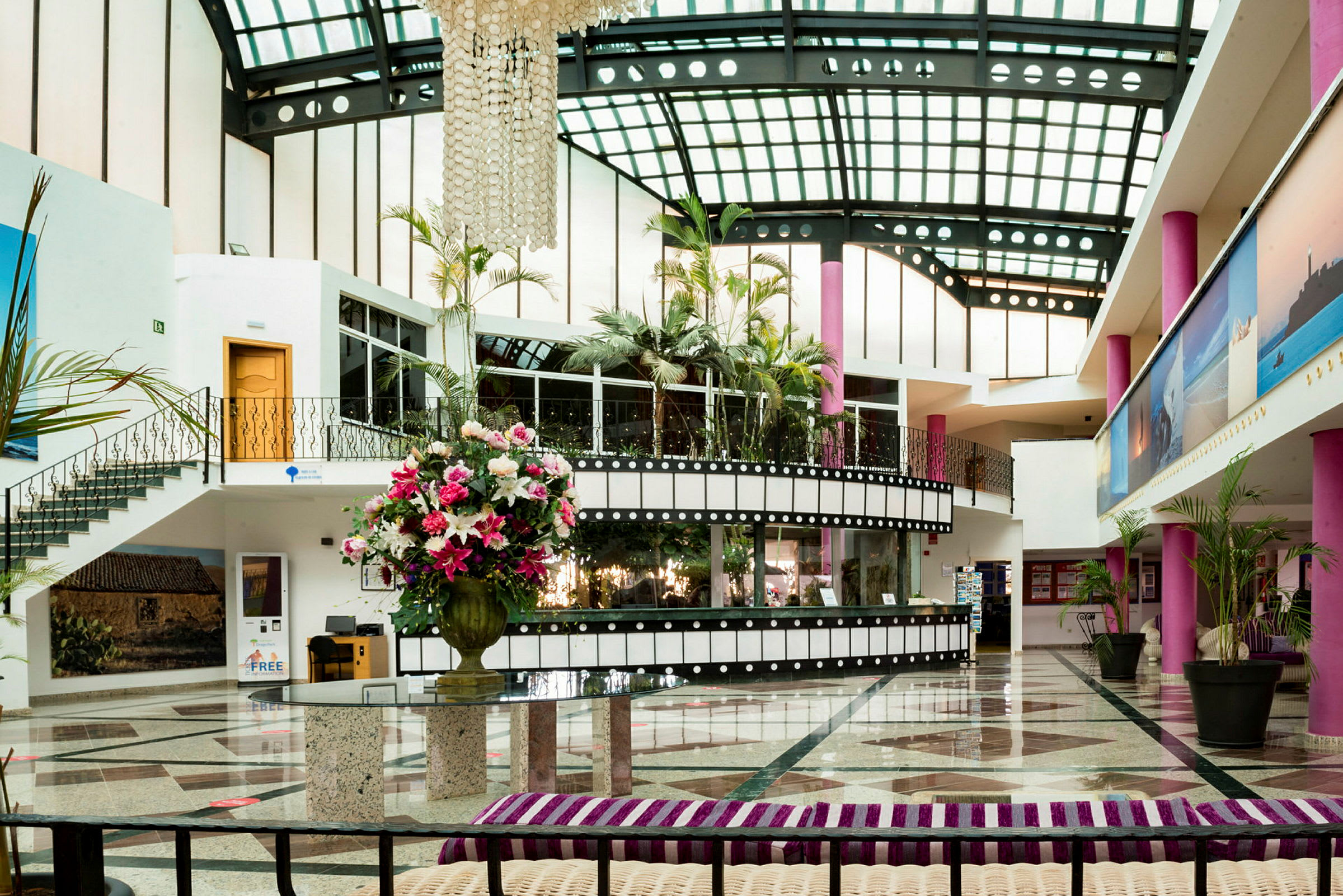 Interiör av en hotellobby med glastak, rosa pelare och en stor vas med färgglada blommor. (AI-genererad text)