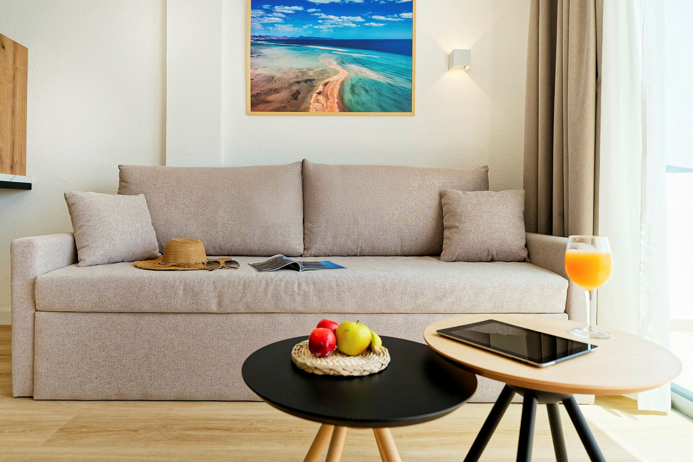 Ett vardagsrum med en beige soffa, en hatt och solglasögon på soffan, samt två små bord med frukt, en surfplatta och ett glas juice. (AI-genererad text)