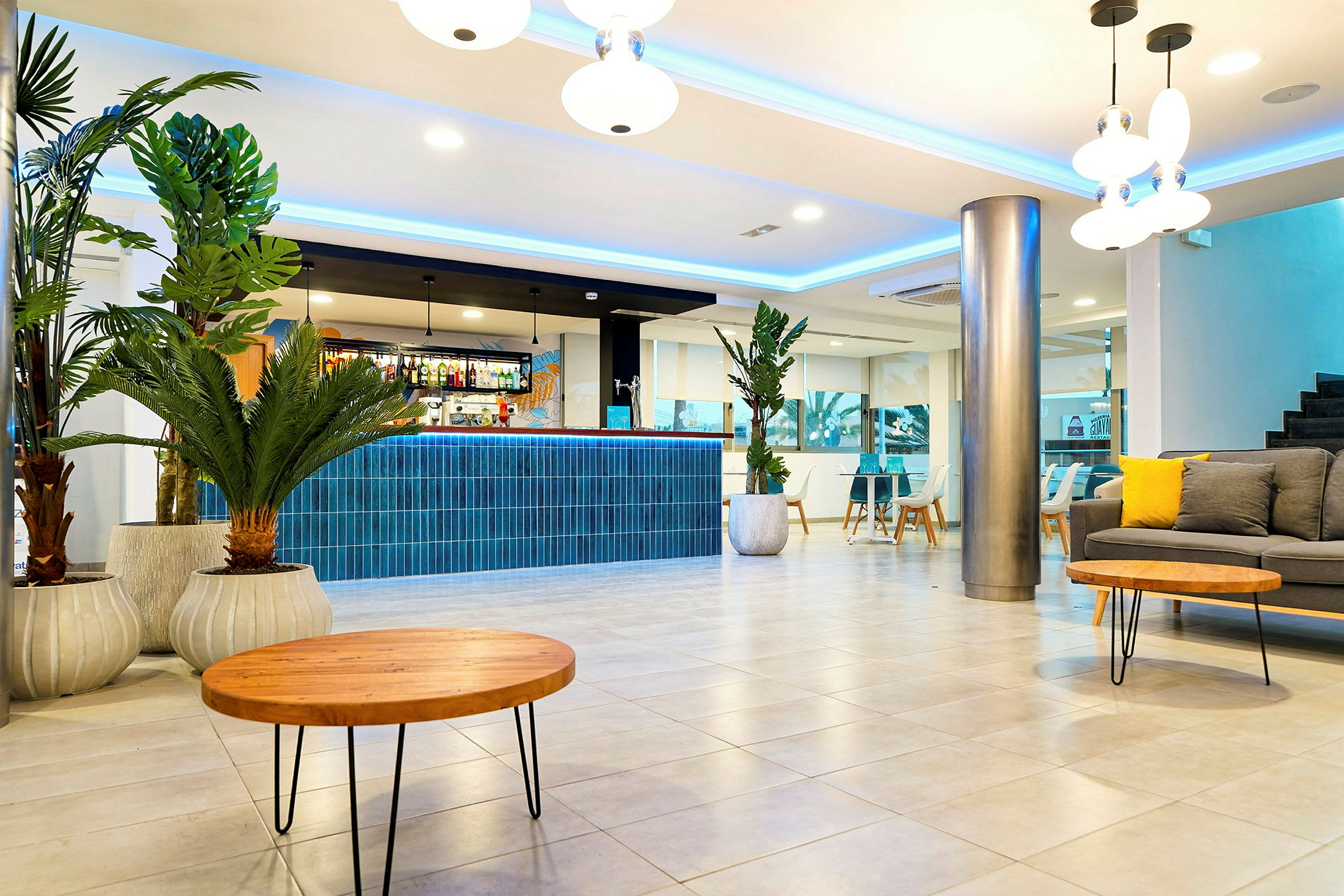 Ett modernt hotellrum med en reception dekorerad med blå kakel, några gröna växter i krukor, en grå soffa med gula kuddar och runda träbord. (AI-genererad text)