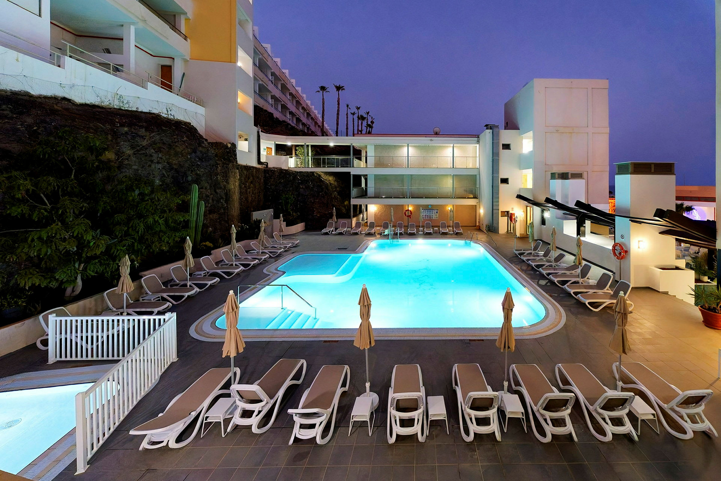 Bilden visar en upplyst hotellpool med omgivande solstolar och parasoller på kvällen. (AI-genererad text)