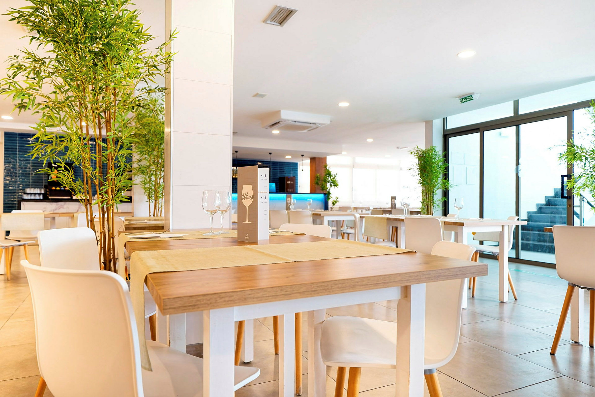 Ett modernt och ljust inrett restaurangutrymme med träbord och vita stolar samt dekorativa växter placerade i hörnen. (AI-genererad text)