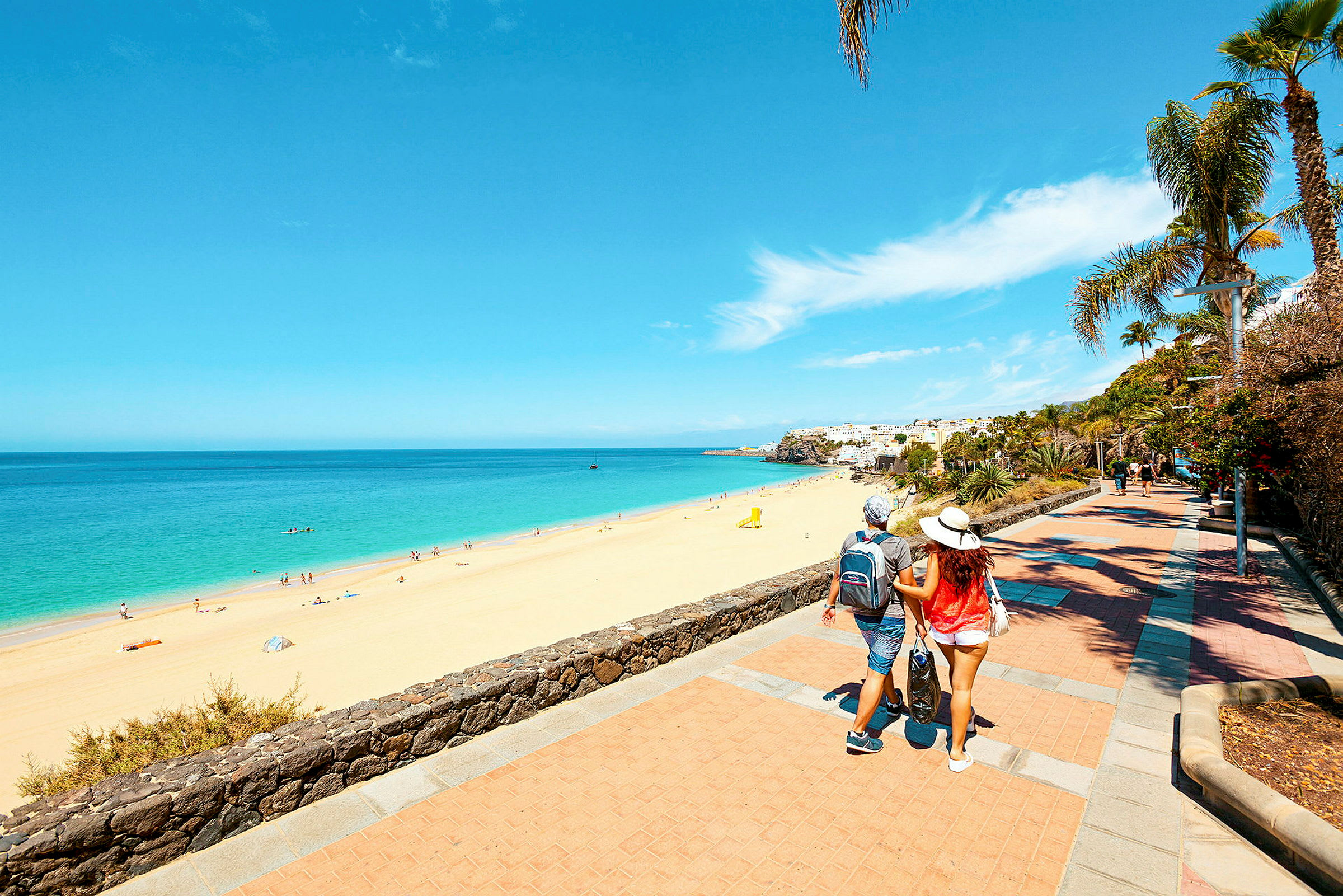 En strand med gyllen sand och klarblått hav, omgiven av en promenad med palmer och annat grönska. En person går längs promenaden. (AI-genererad text)