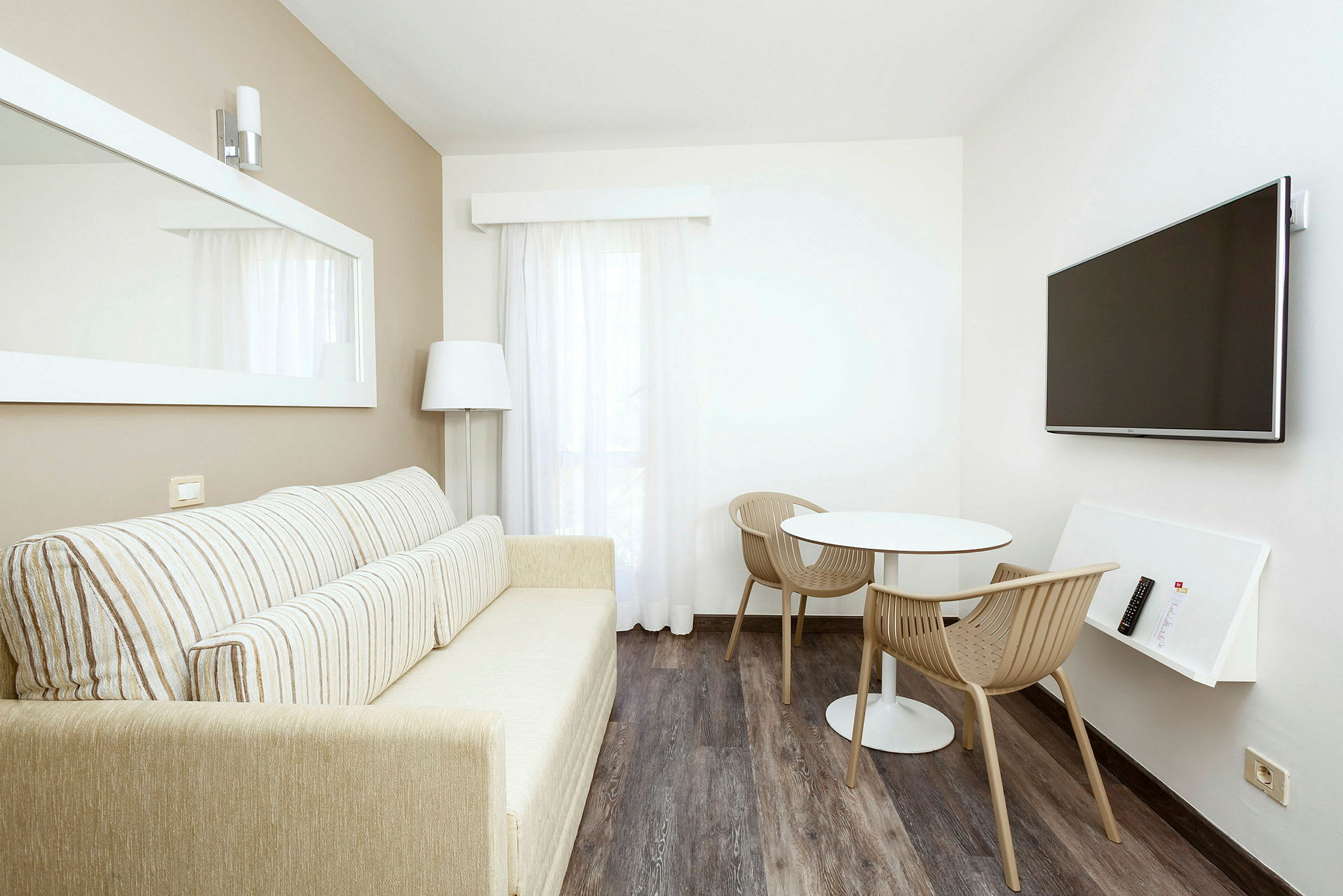 Ett ljust vardagsrum med en soffa, ett litet bord med två stolar och en väggmonterad TV. (AI-genererad text)