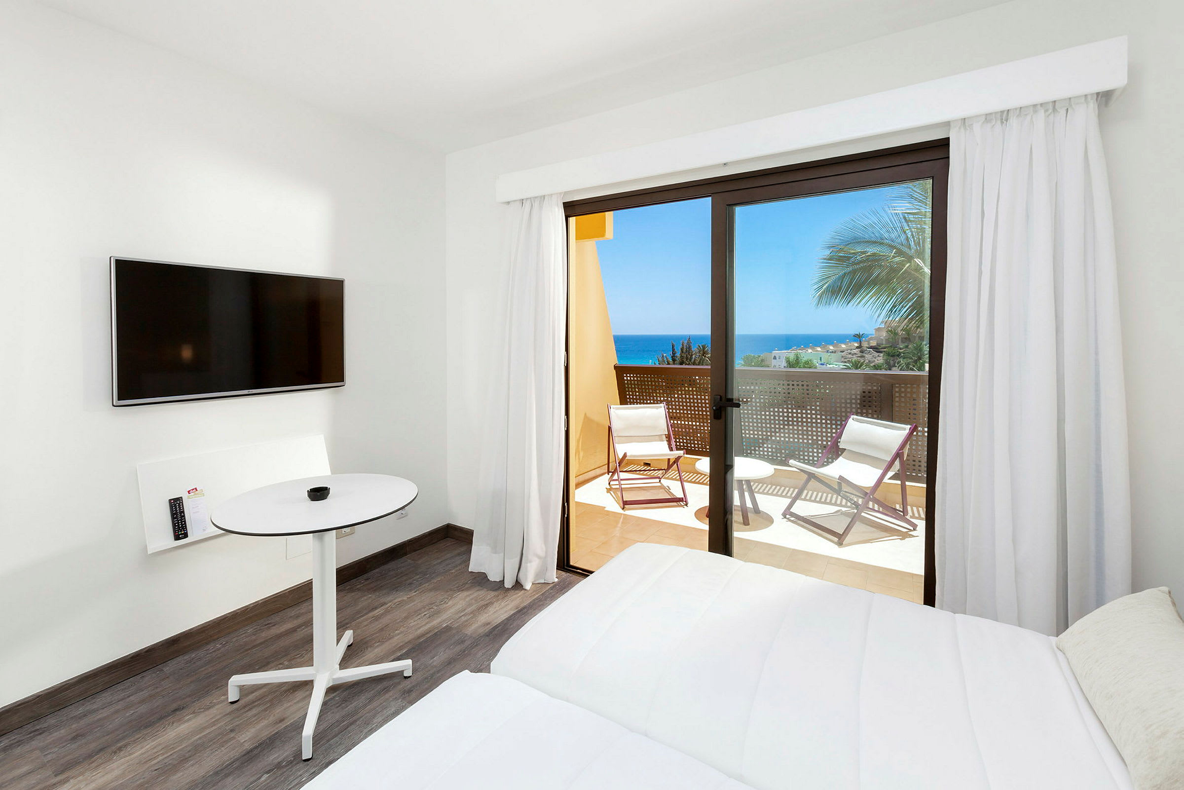 Ett hotellrum med en vit säng, en väggmonterad TV, ett runt bord och en balkong med utsikt över havet. (AI-genererad text)
