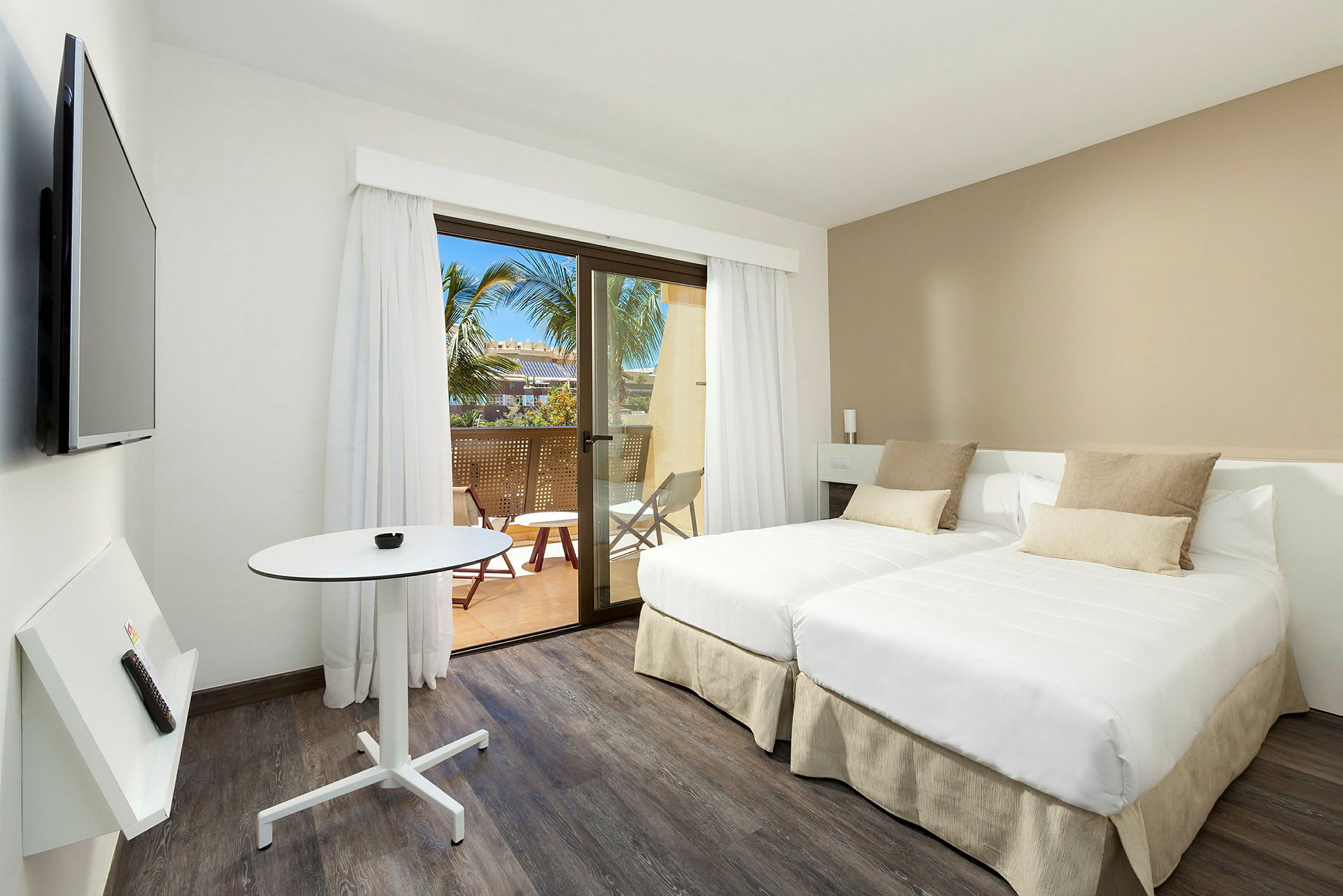 Ett modernt hotellrum med två enkelsängar, ett mindre bord och en väggmonterad TV. Rummet har ljusa färger och ett fönster som leder ut till en balkong med utsikt mot palmer. (AI-genererad text)