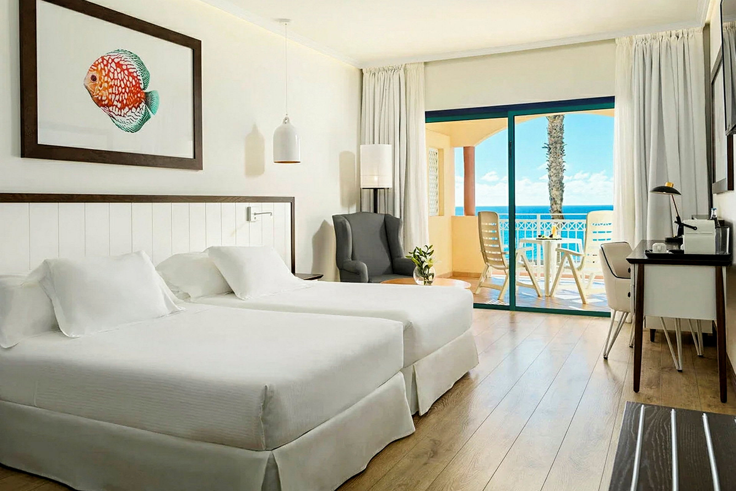 Ett hotellrum med två sängar, en fåtölj, ett skrivbord och en balkong med utsikt mot havet. (AI-genererad text)