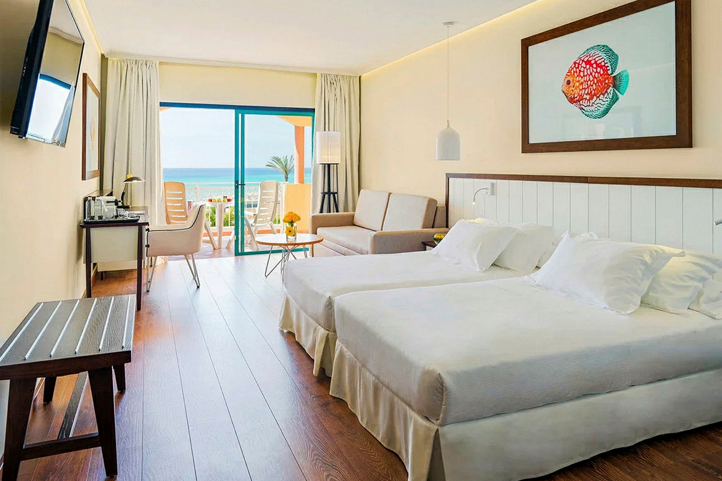 Ett hotellrum med två bäddar, en soffa och ett bord. Rummet har en altandörr som leder till en balkong med havsutsikt. (AI-genererad text)
