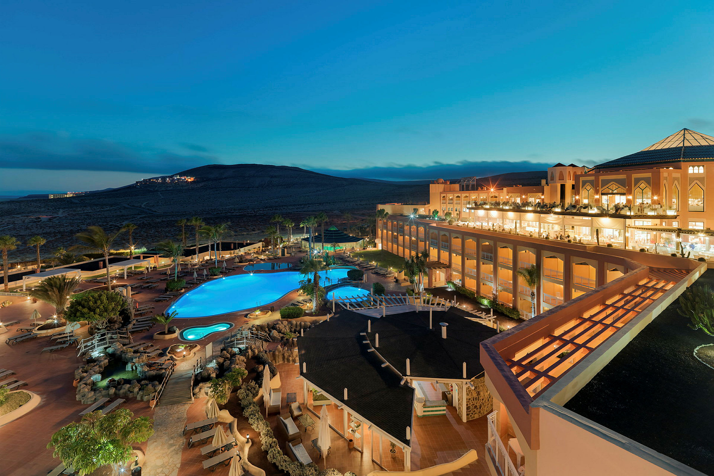 En vy över ett hotellkomplex med pool och omgivande landskap i kvällsljus. (AI-genererad text)