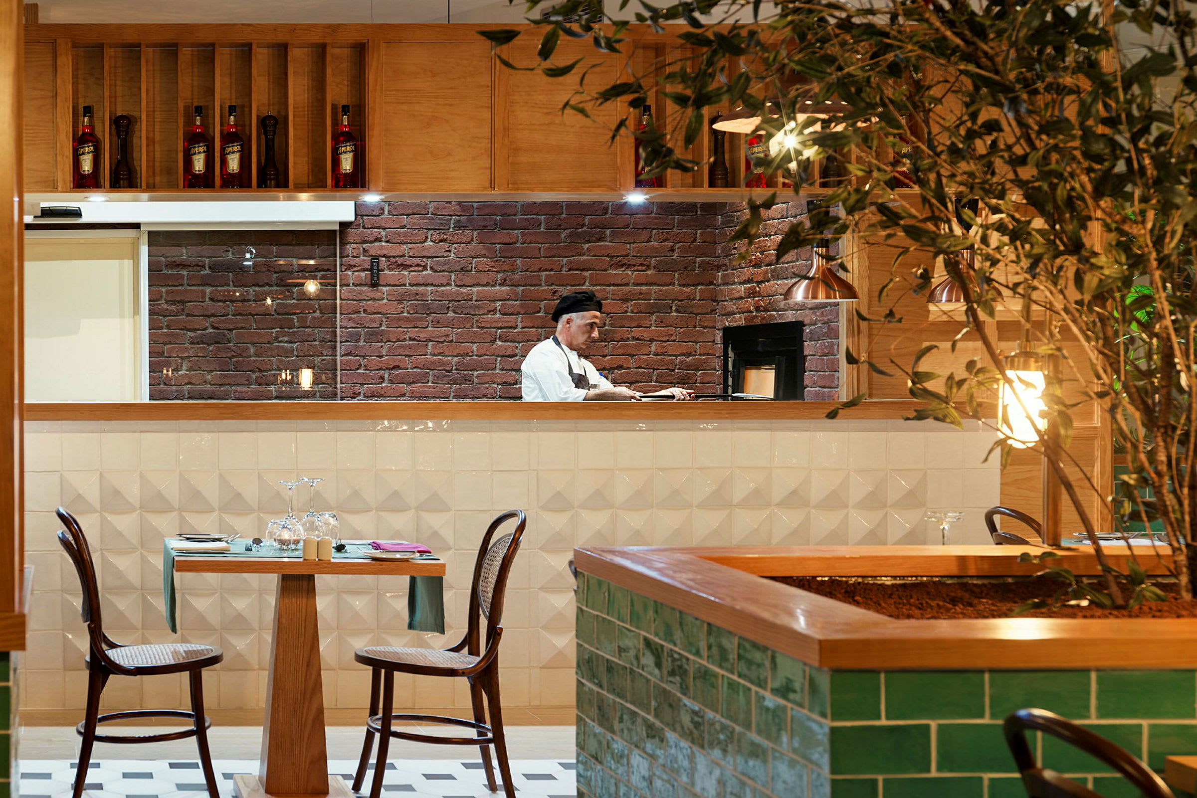 En restaurang med ett öppet kök där en person arbetar. Ett bord med två stolar står framför köket, och en växt är synlig i förgrunden. (AI-genererad text)