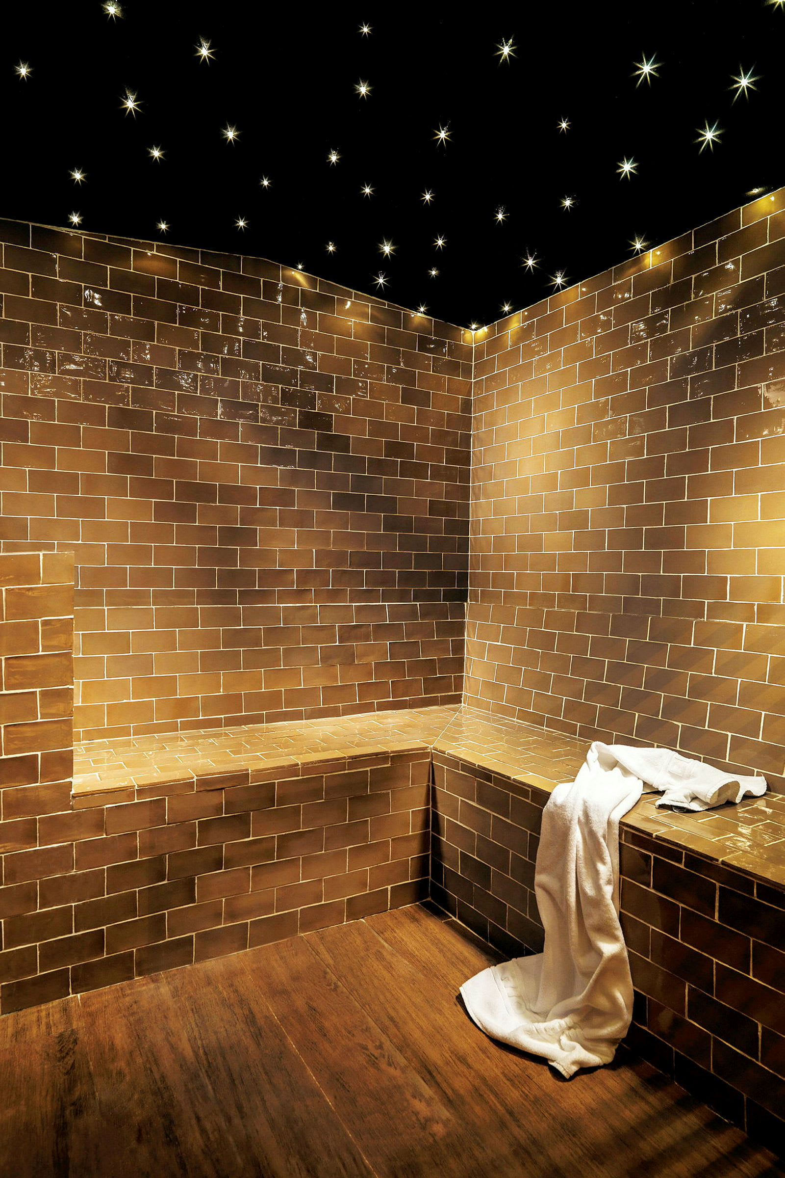 Ett spa med bruna kakelklädda väggar och sittplatser, ett vitt handduk drapera över kanten, och taket dekorerat med stjärnformade lampor. (AI-genererad text)