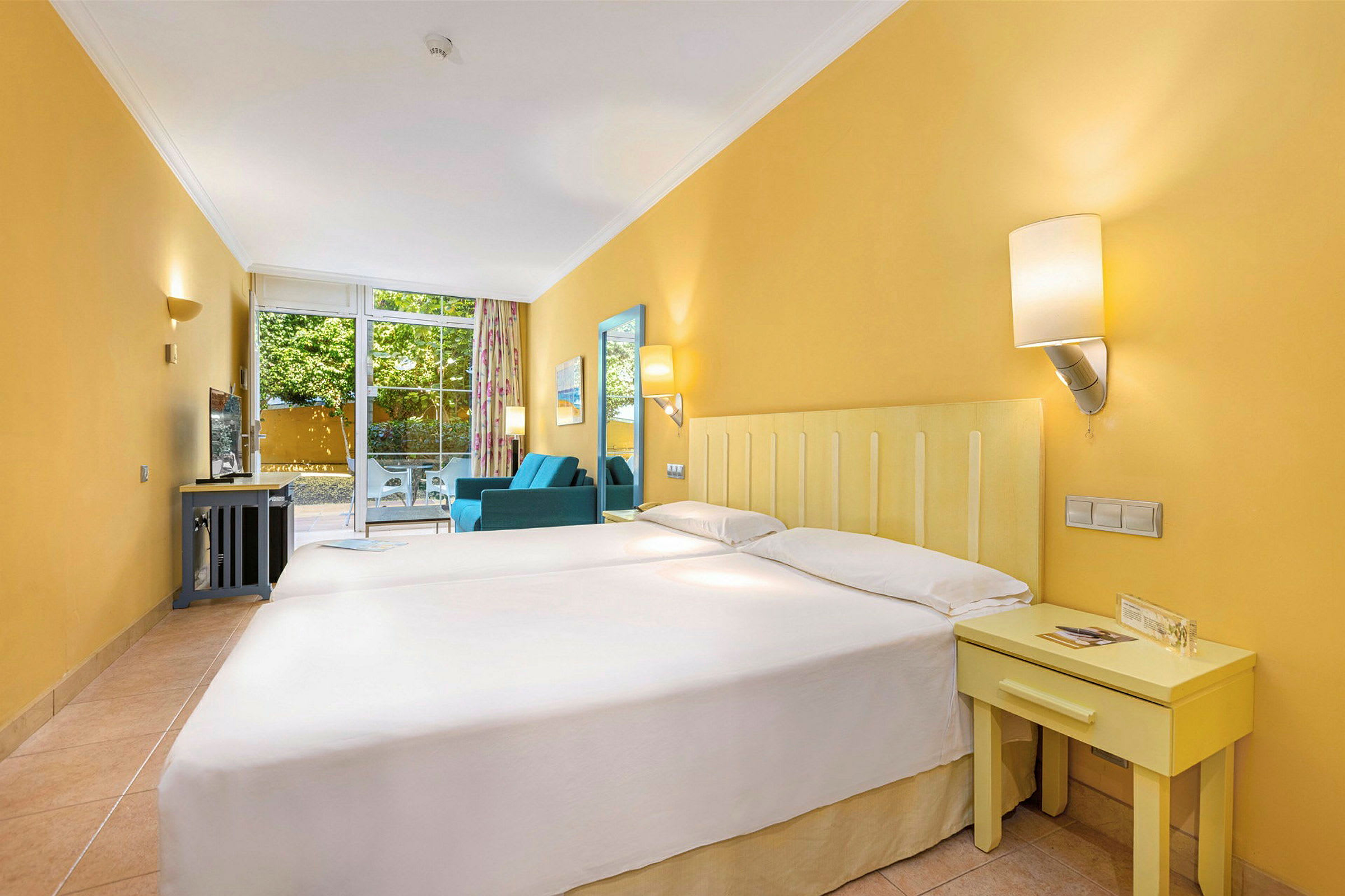 Ett hotellrum med två sängar, en soffa, bord och en balkongdörr som leder ut till en terrass med utsikt över träd och buskar. (AI-genererad text)