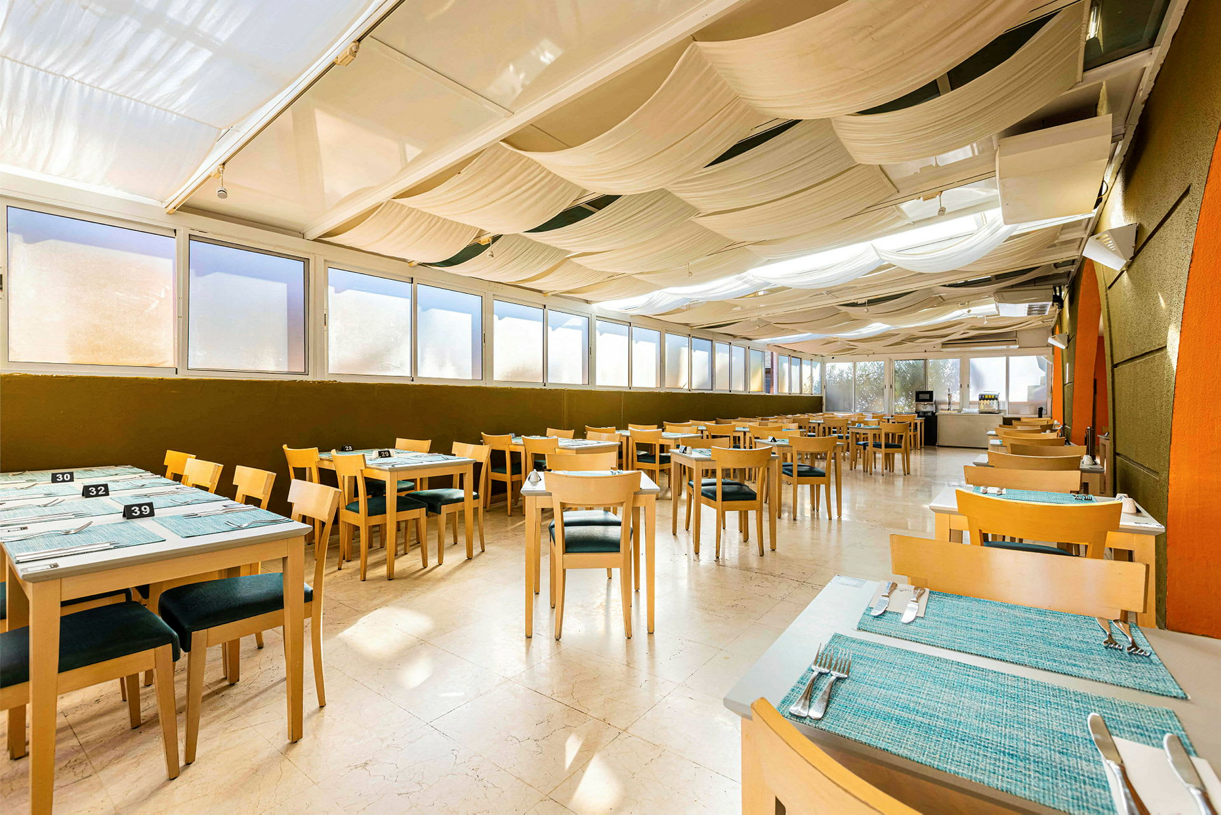 Bild på en inomhusrestaurang med träbord och stolar, blå bordmattor och dagsljus från stora fönster. (AI-genererad text)