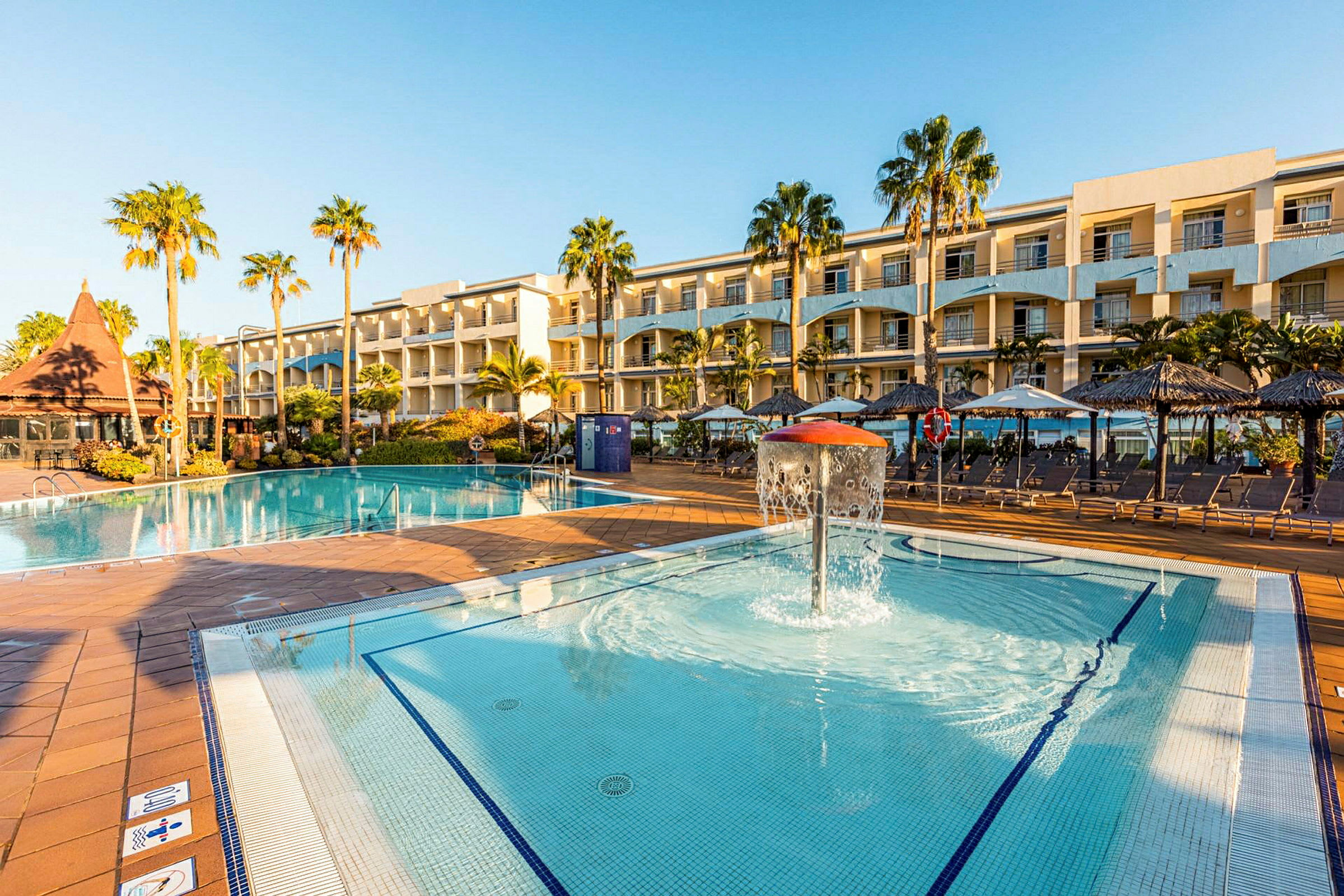 En stor hotellpool med omgivande solstolar och palmer, samt en mindre barnpool med vattenfontän framför hotellets fasad. (AI-genererad text)