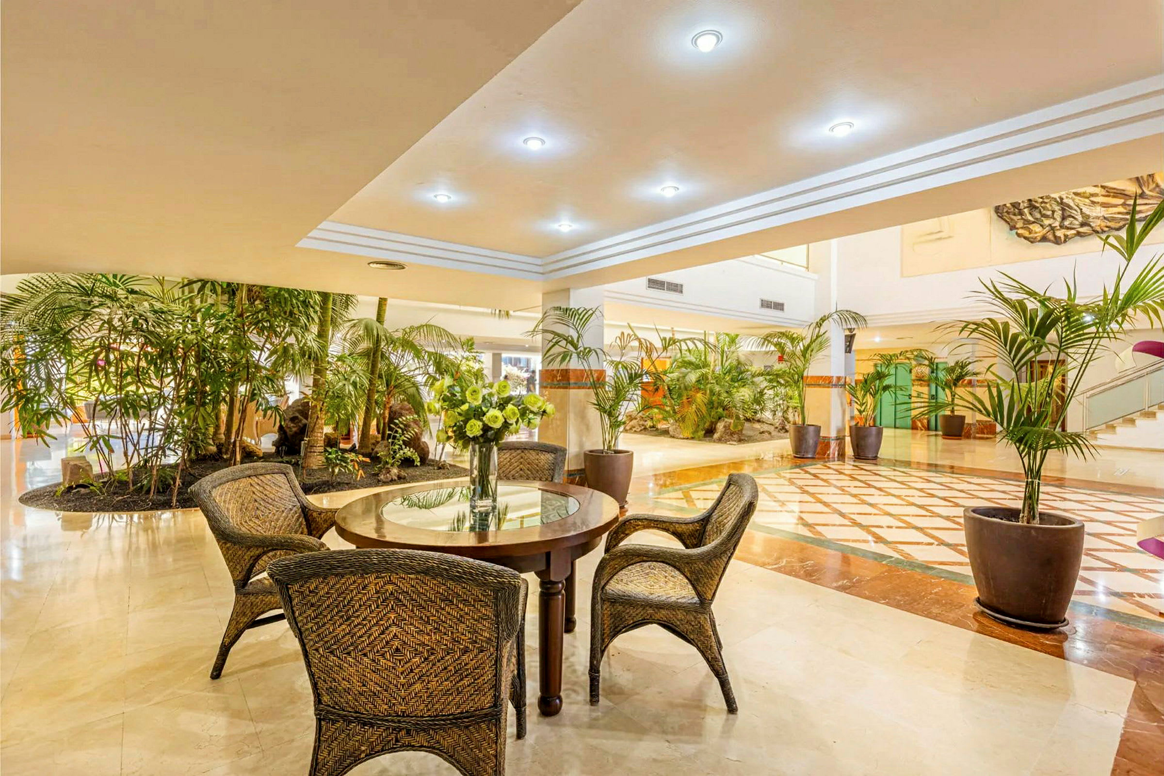 Ett ljust hotellatrium med sittmöbler, ett runt bord med blomvas och många gröna växter. (AI-genererad text)