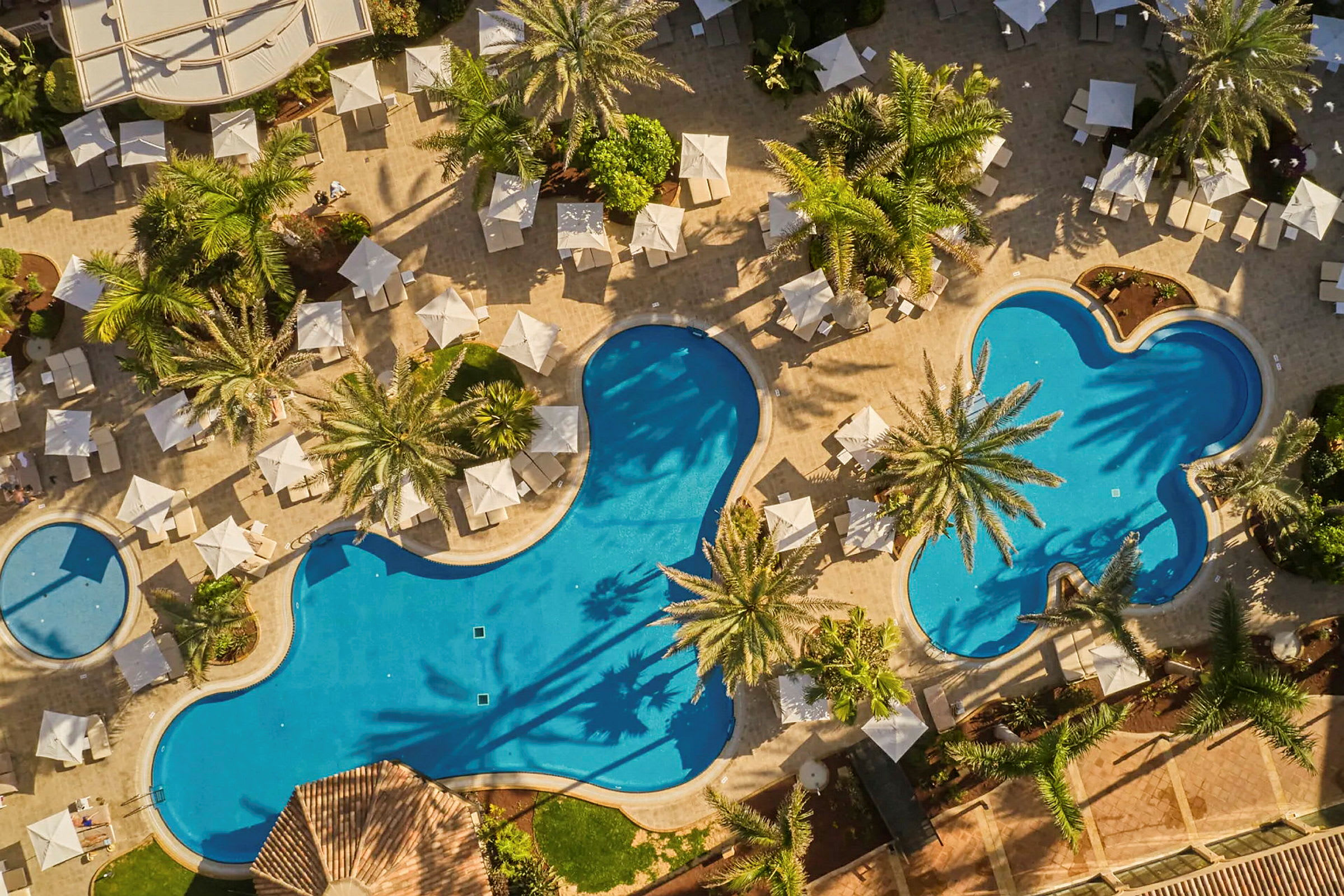 Flygfoto av en gård med tre swimmingpooler och områden med palmer, solstolar och parasoller. (AI-genererad text)