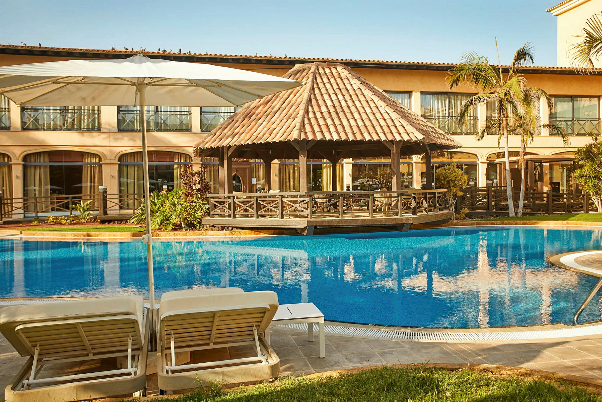 Hotellområde med pool, solsängar och parasoll framför en byggnad med stora fönster och en träpaviljong. (AI-genererad text)