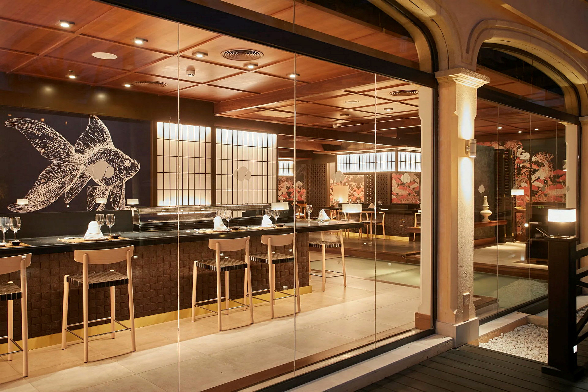En elegant restaurang med en bar och sittplatser. Det finns en stor väggdekoration med ett fiskmotiv och belysning som skapar en varm atmosfär. (AI-genererad text)