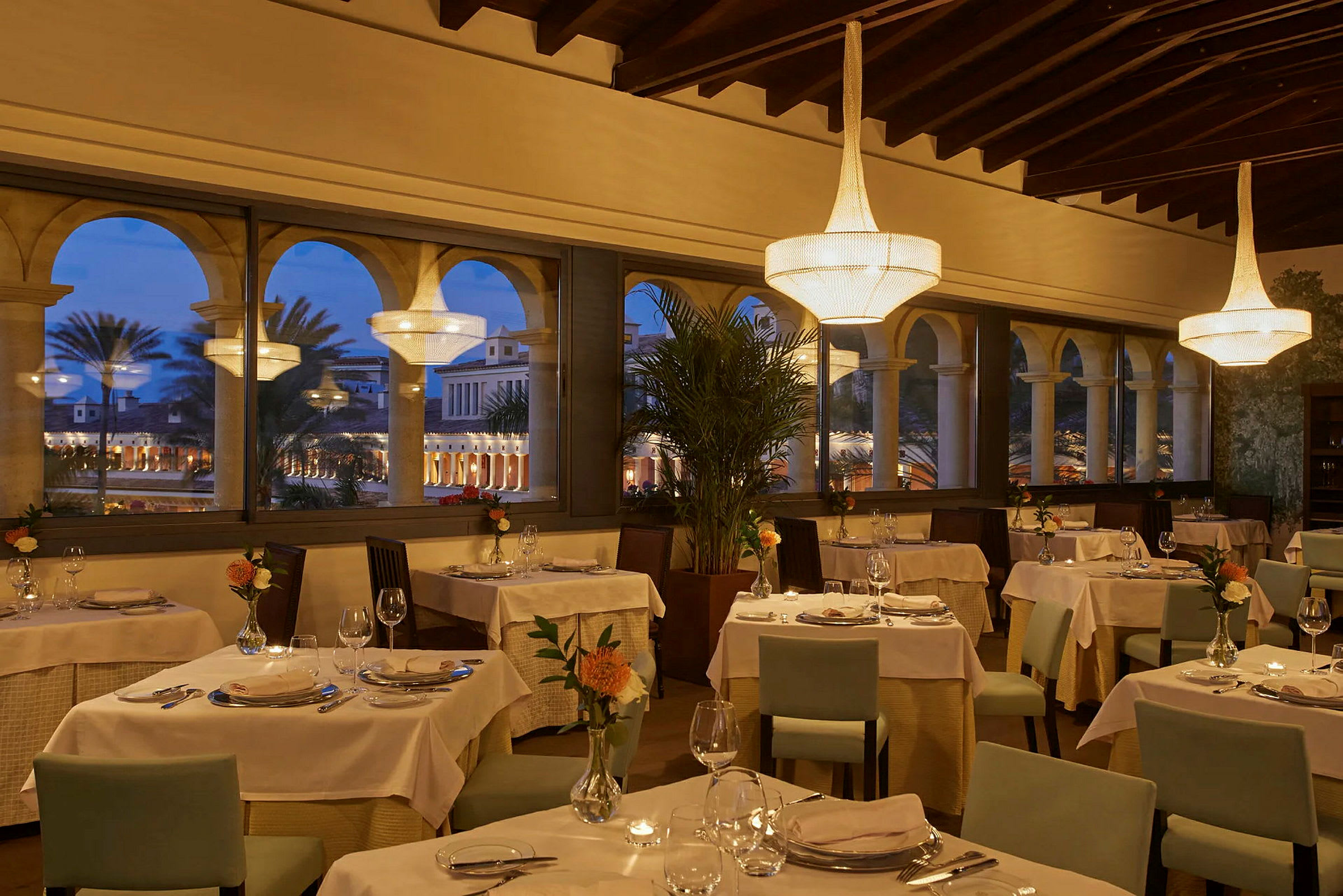 Interiör av en elegant restaurang med dukade bord och stora fönster mot en kvällsbelyst innergård. (AI-genererad text)