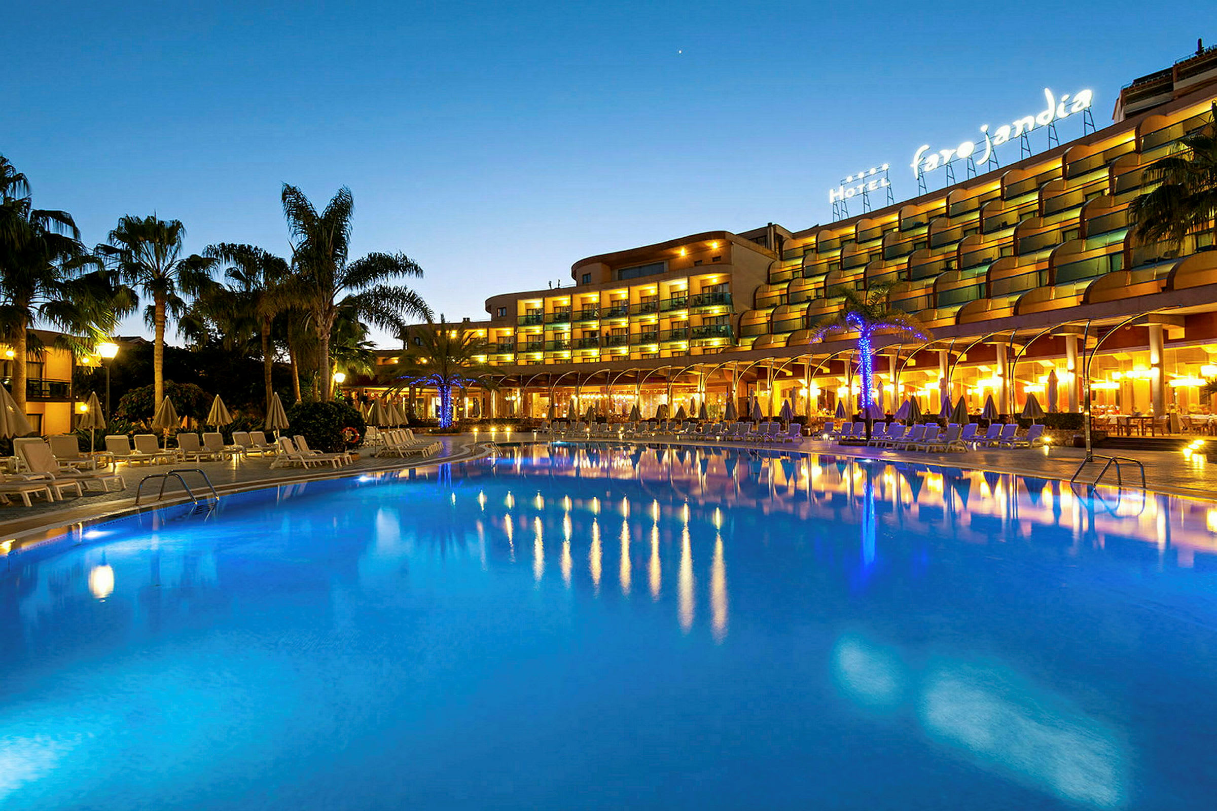 Upplyst hotell vid en stor uppvärmd pool omgiven av solstolar och palmer på kvällen. (AI-genererad text)