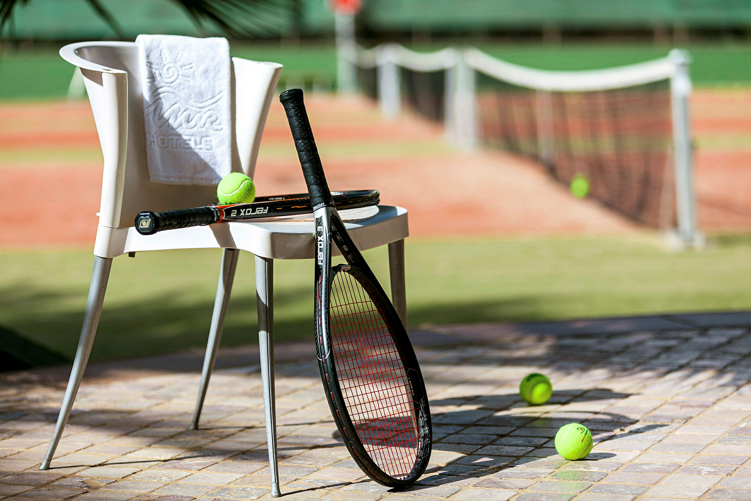 En stol med en handduk och två tennisracketar placerad vid sidan av en tennisbana, med tennisbollar på marken. (AI-genererad text)