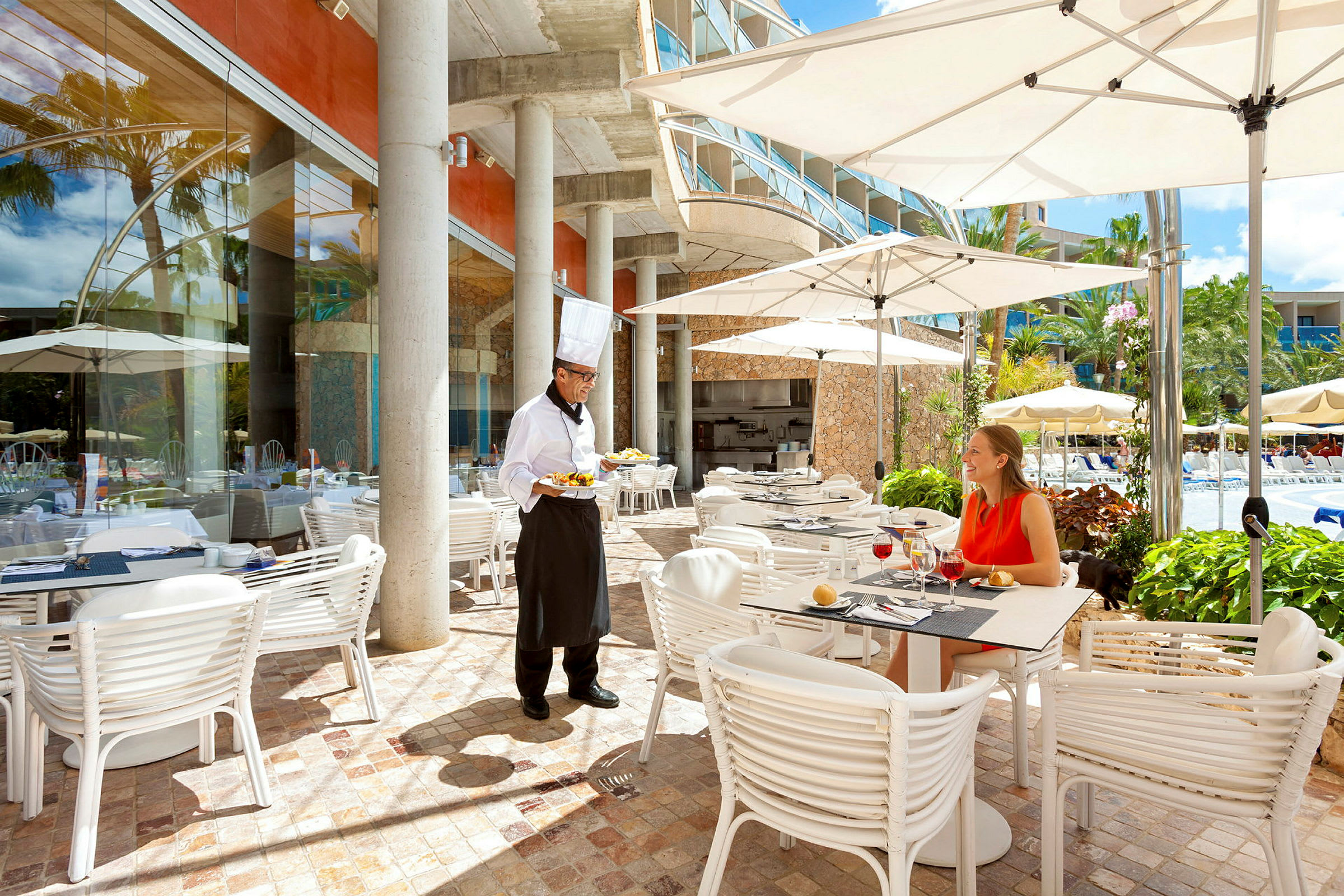 En restaurangterrass med bord, stolar och parasoll bredvid en pool. En servitör serverar mat till en gäst. (AI-genererad text)