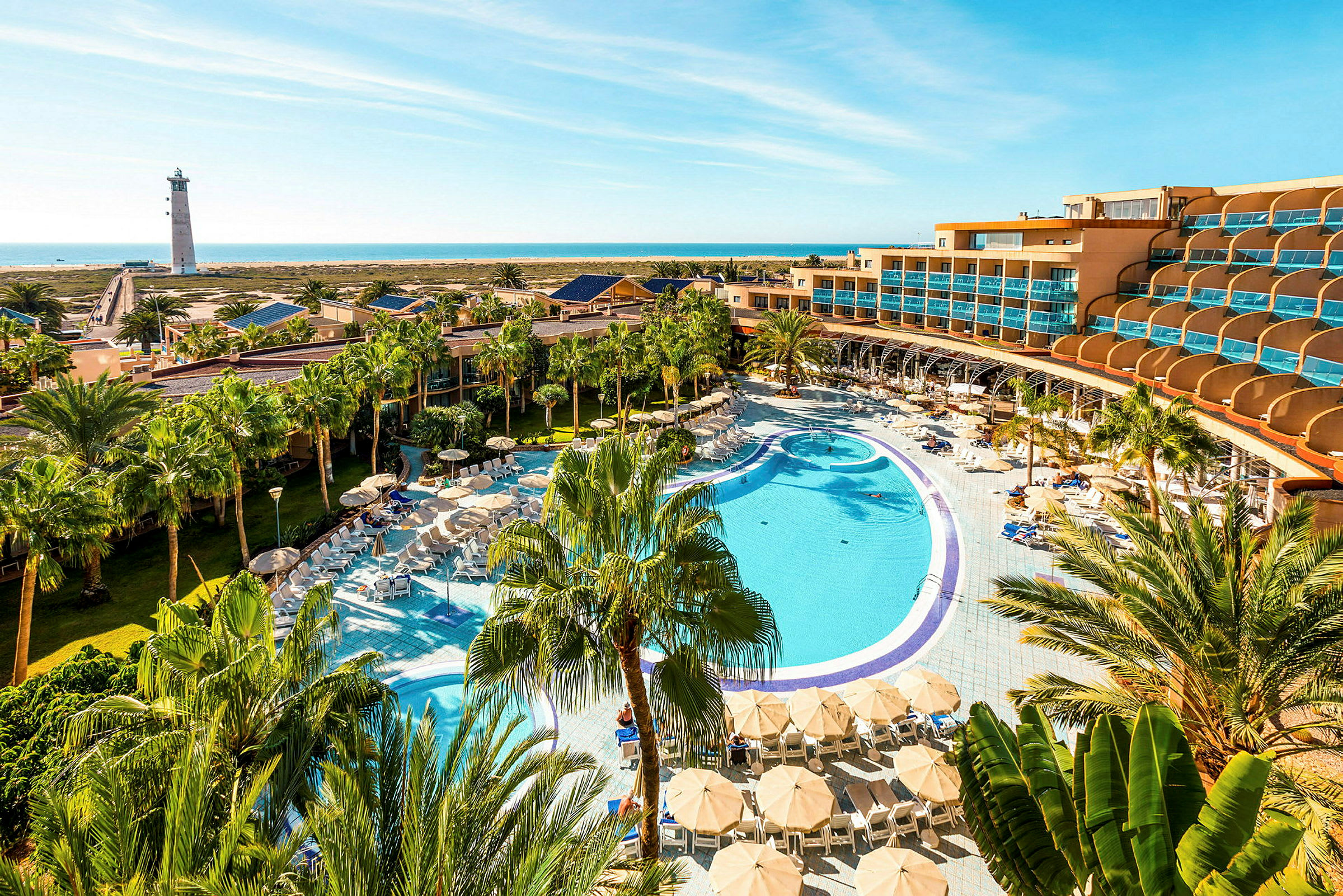 En hotellanläggning med en stor pool, solstolar och parasoller omgiven av palmer, med havet och en fyr i bakgrunden. (AI-genererad text)