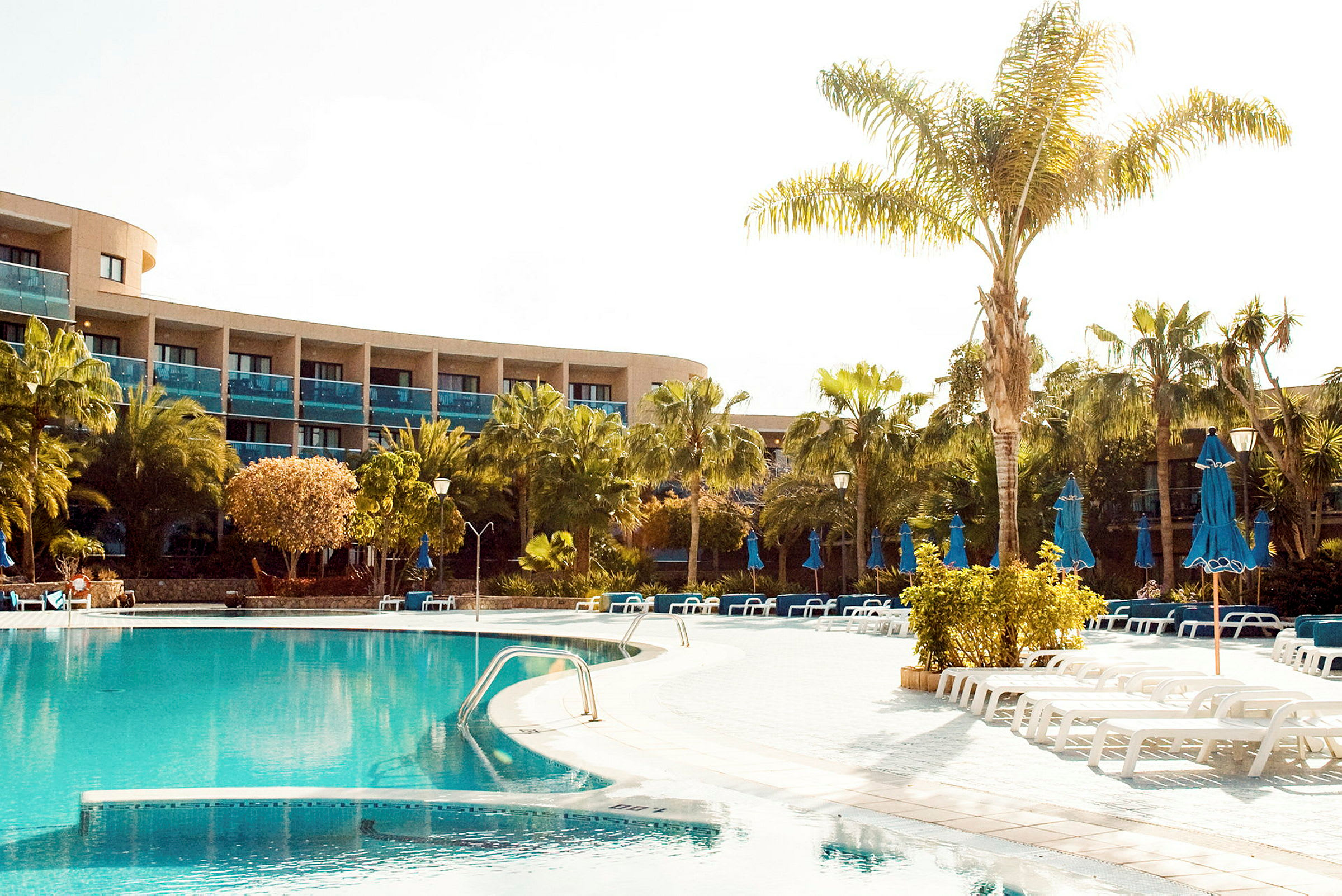 En hotellpool med solsängar och parasoller samt omgivande grönska och palmer. (AI-genererad text)