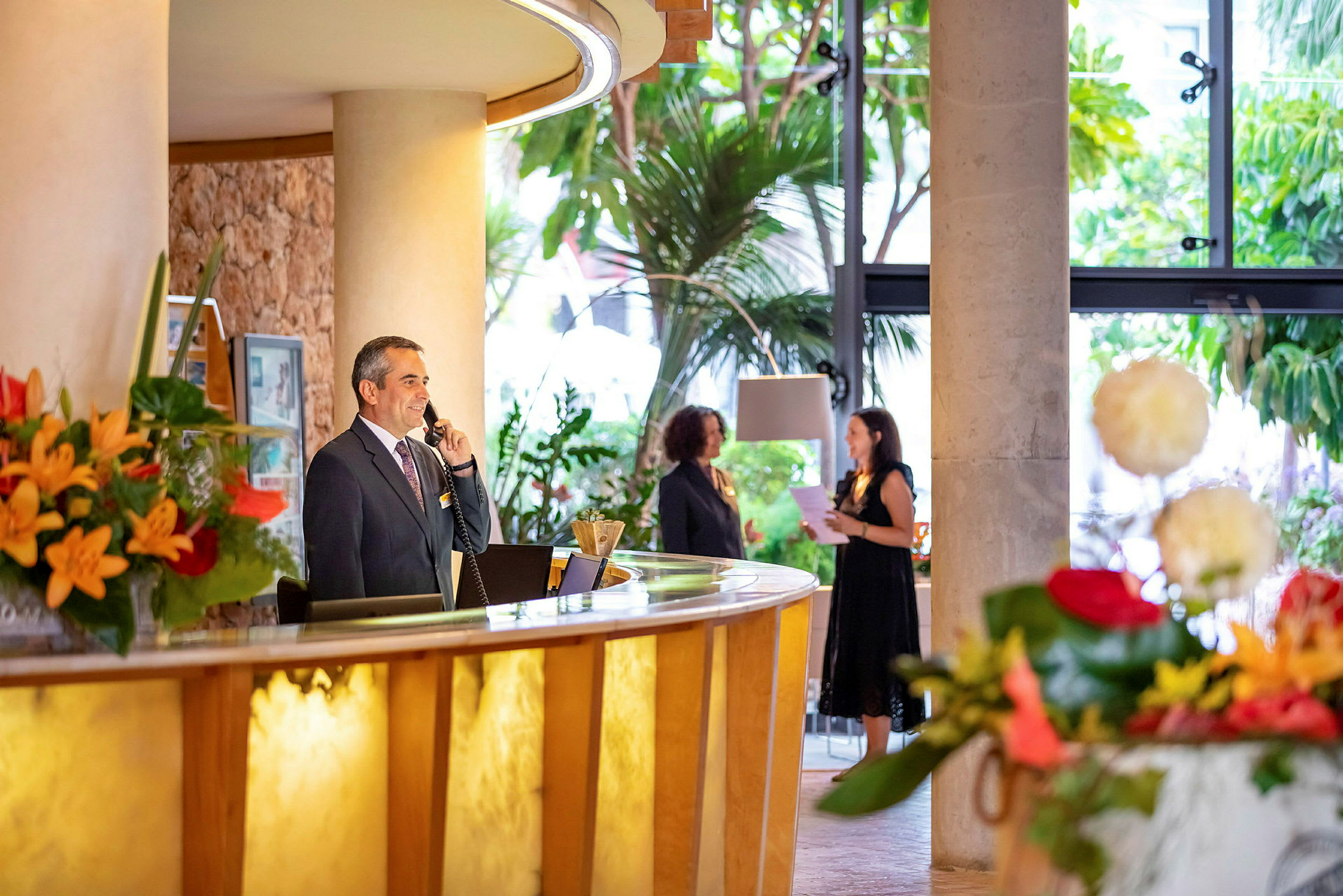 En hotellreception med blommor, en receptionist vid disken och gäster i bakgrunden. (AI-genererad text)