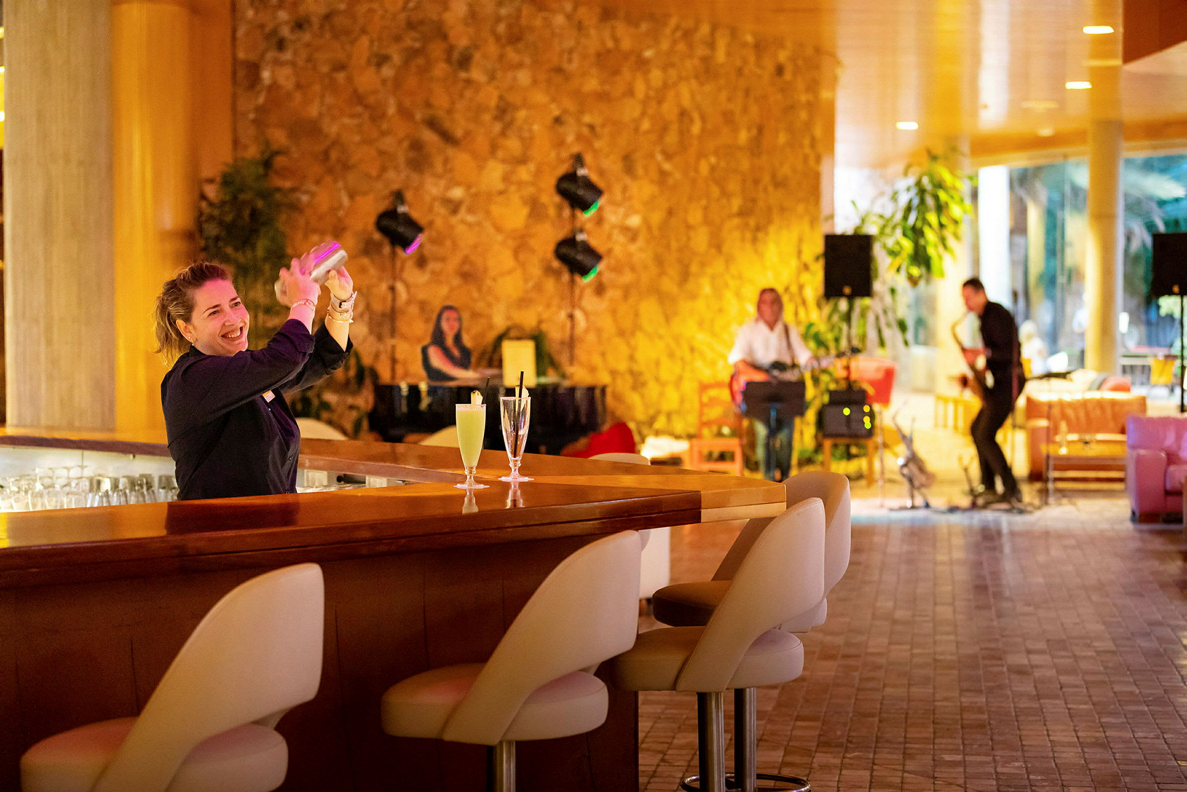 En bartender tillreder en drink vid bardisken. I bakgrunden syns musiker som spelar instrument i en varm och stilfull miljö. (AI-genererad text)