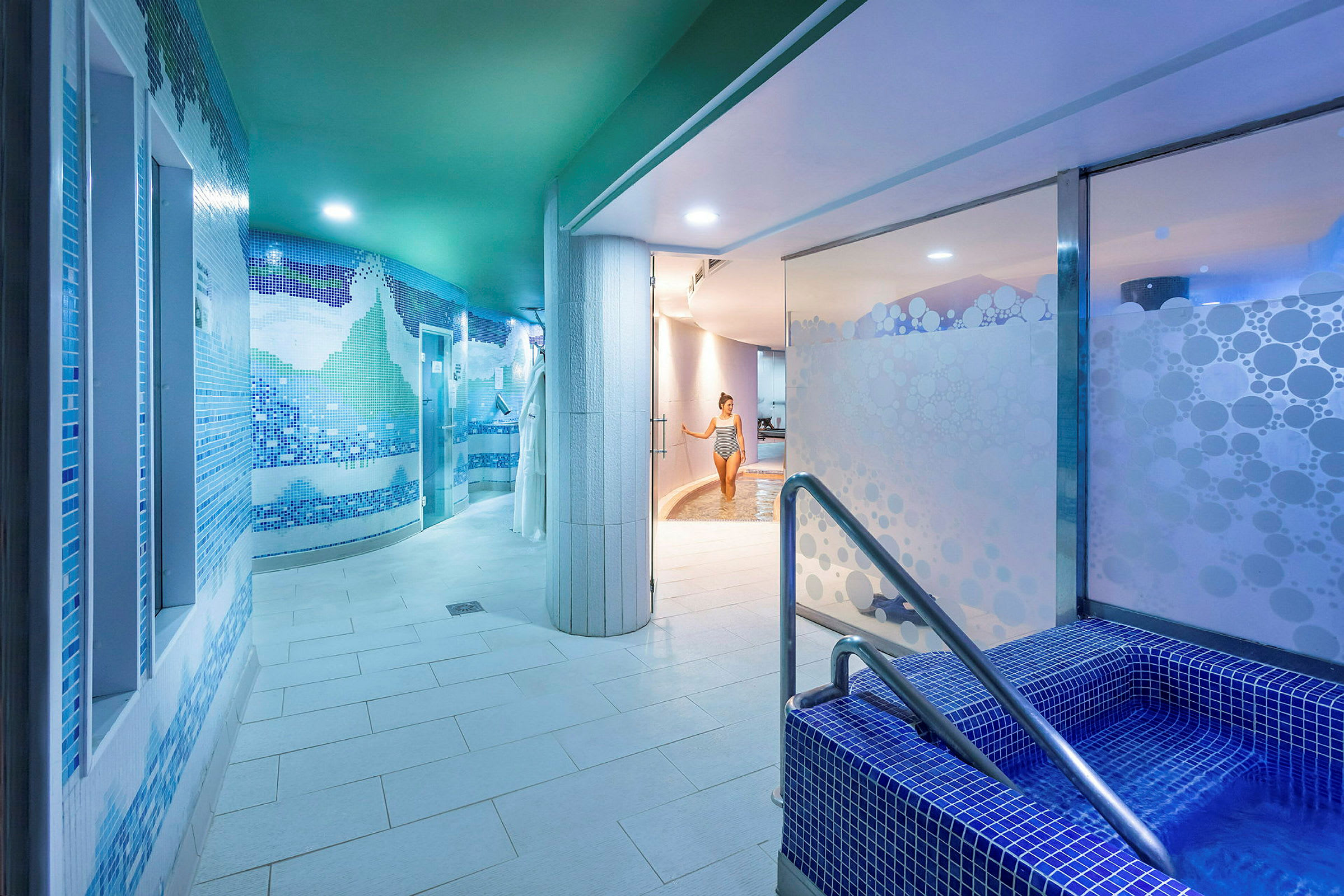 Ett inomhusspa med blå mosaikväggar, en mindre pool med trappor i förgrunden och en person som går mot en större bassäng i bakgrunden. (AI-genererad text)