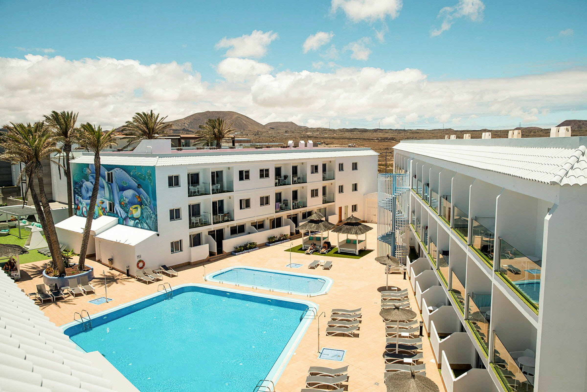 Bilden visar ett hotellkomplex med vita byggnader. En stor pool och en mindre barnpool är omgivna av solstolar och parasoller. Bakgrunden består av palmer och ett berg under en blå himmel. (AI-genererad text)