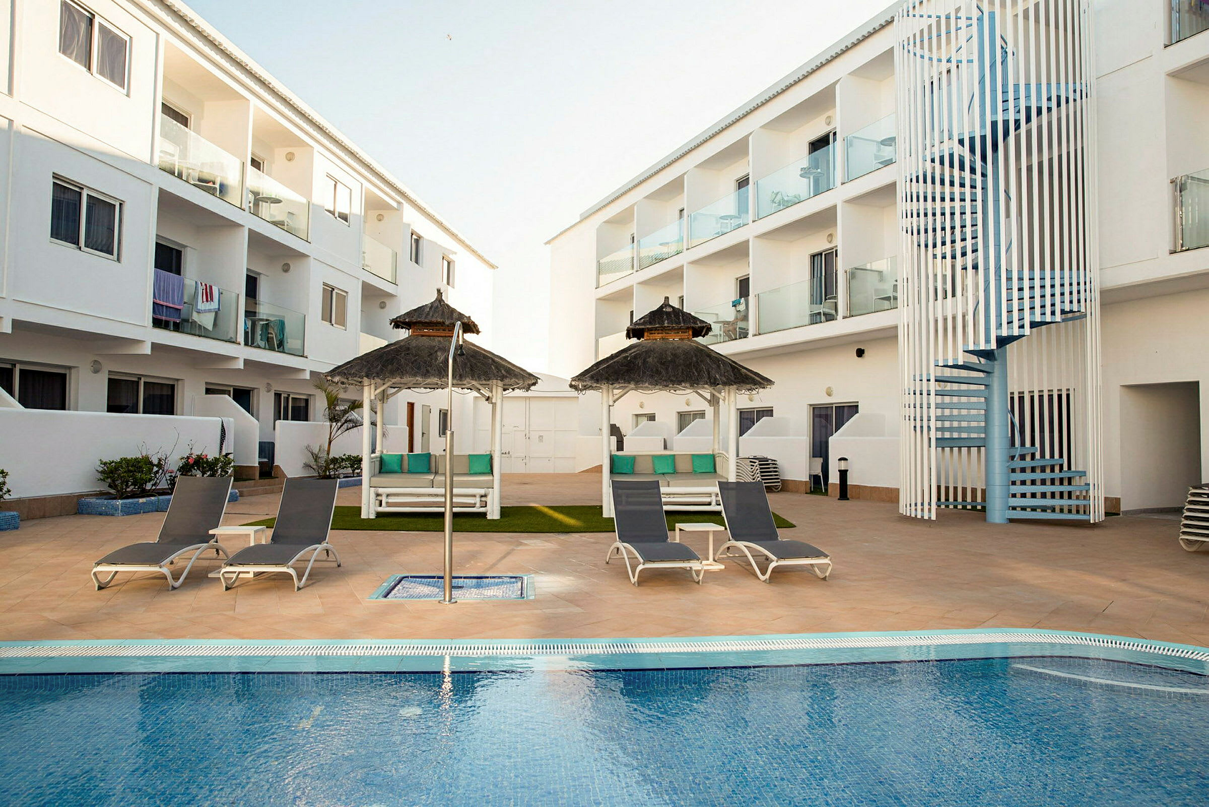 Ett hotellområde med en pool omgiven av solstolar och parasoller. I bakgrunden syns vita byggnader med balkonger. (AI-genererad text)