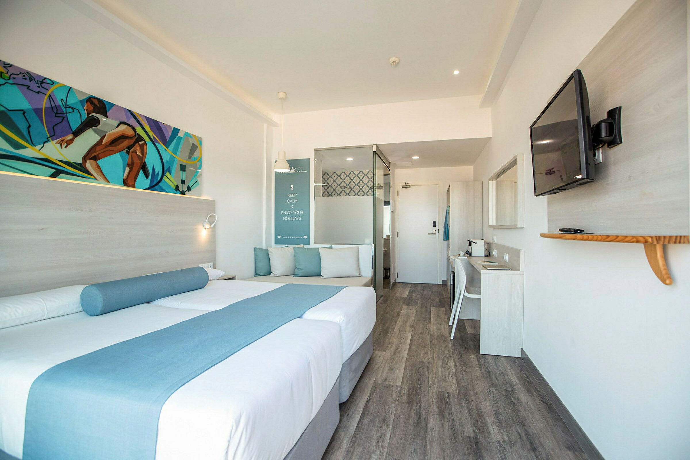 Ett modernt hotellrum med en dubbelsäng dekorerad med blå textilier, en sittplats med kuddar, ett skrivbord, och en väggmonterad TV. (AI-genererad text)