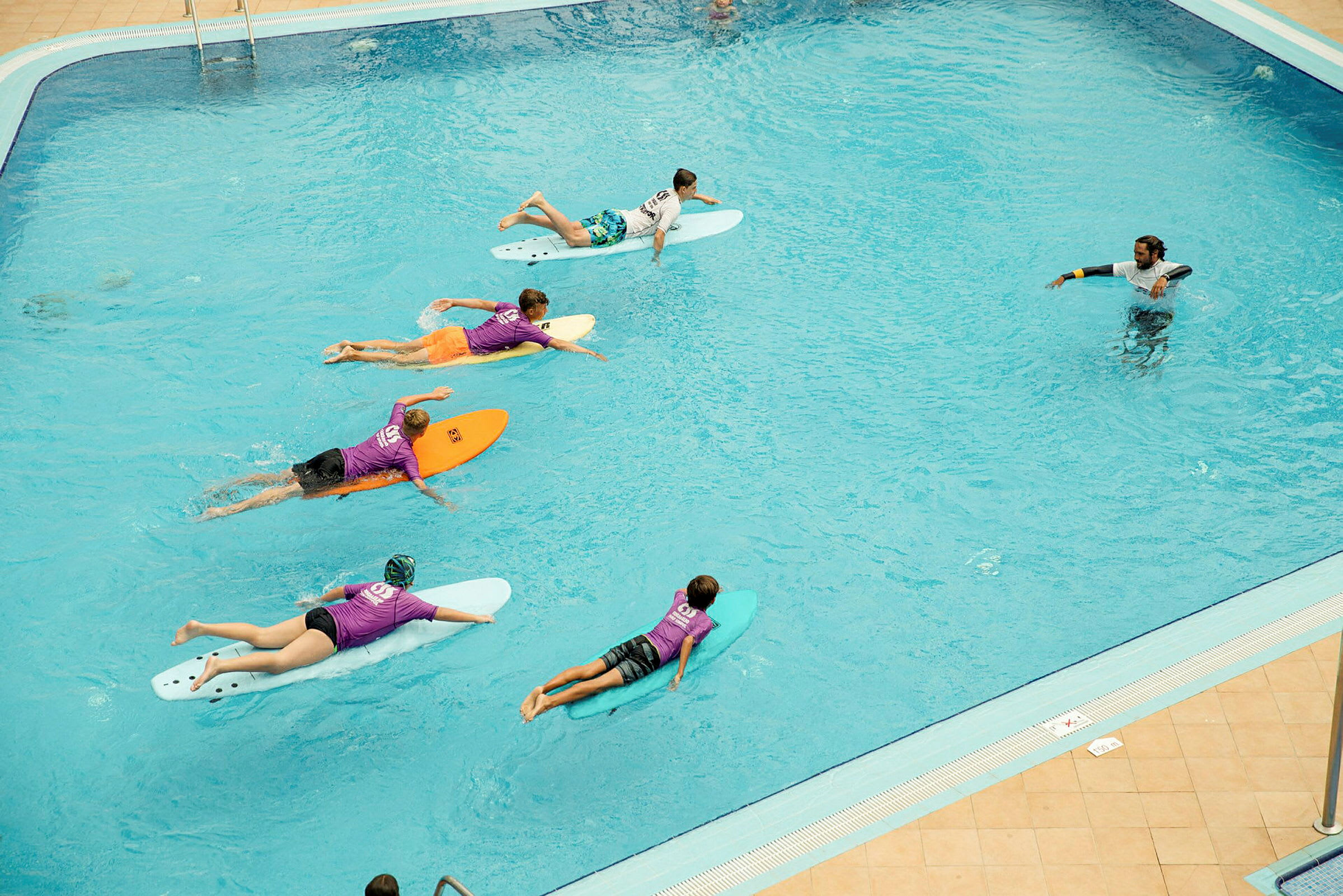 En grupp människor ligger på surfbrädor i en pool, och en instruktör står i vattnet. (AI-genererad text)