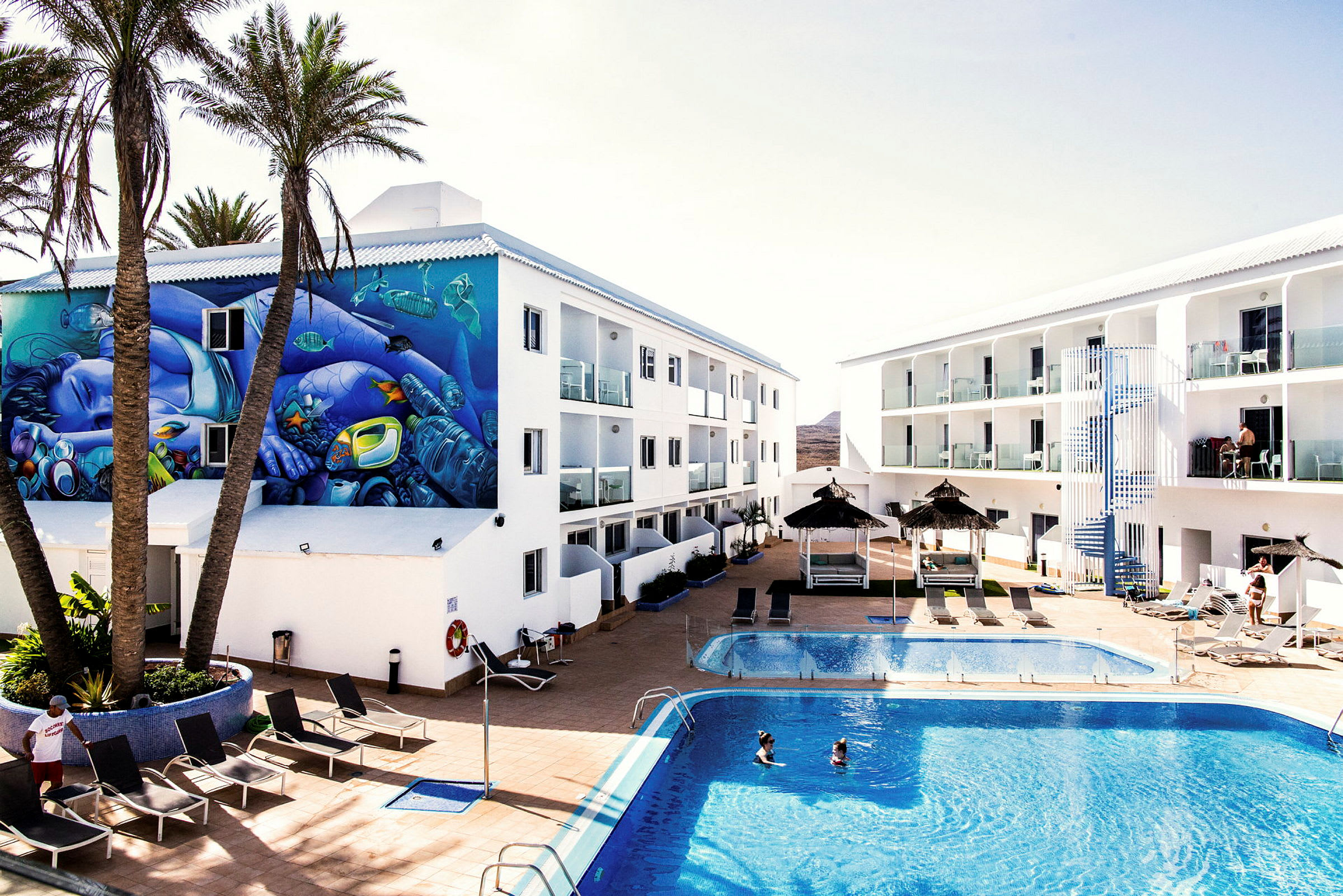 Hotellområde med pool, solstolar, parasoller och två vita byggnader. En vägg har en färgglad muralmålning, och två personer badar i poolen. (AI-genererad text)