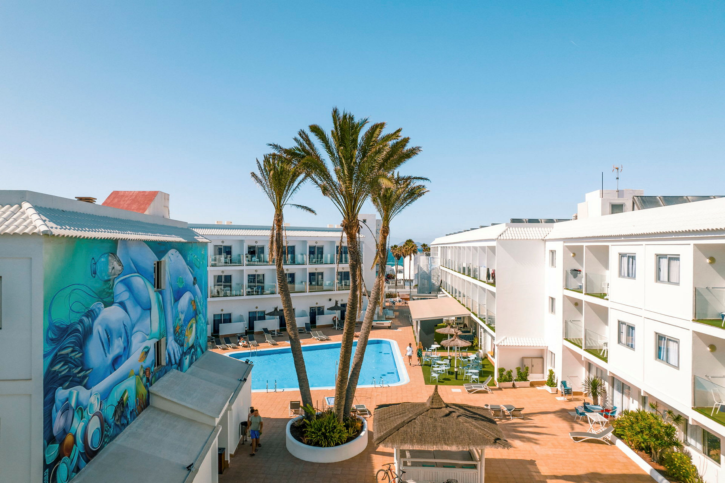 Ett hotellområde med en pool i mitten, omgiven av vita byggnader med balkonger. En väggmålning syns på en av byggnaderna, och det finns palmer och sittplatser vid poolen. (AI-genererad text)